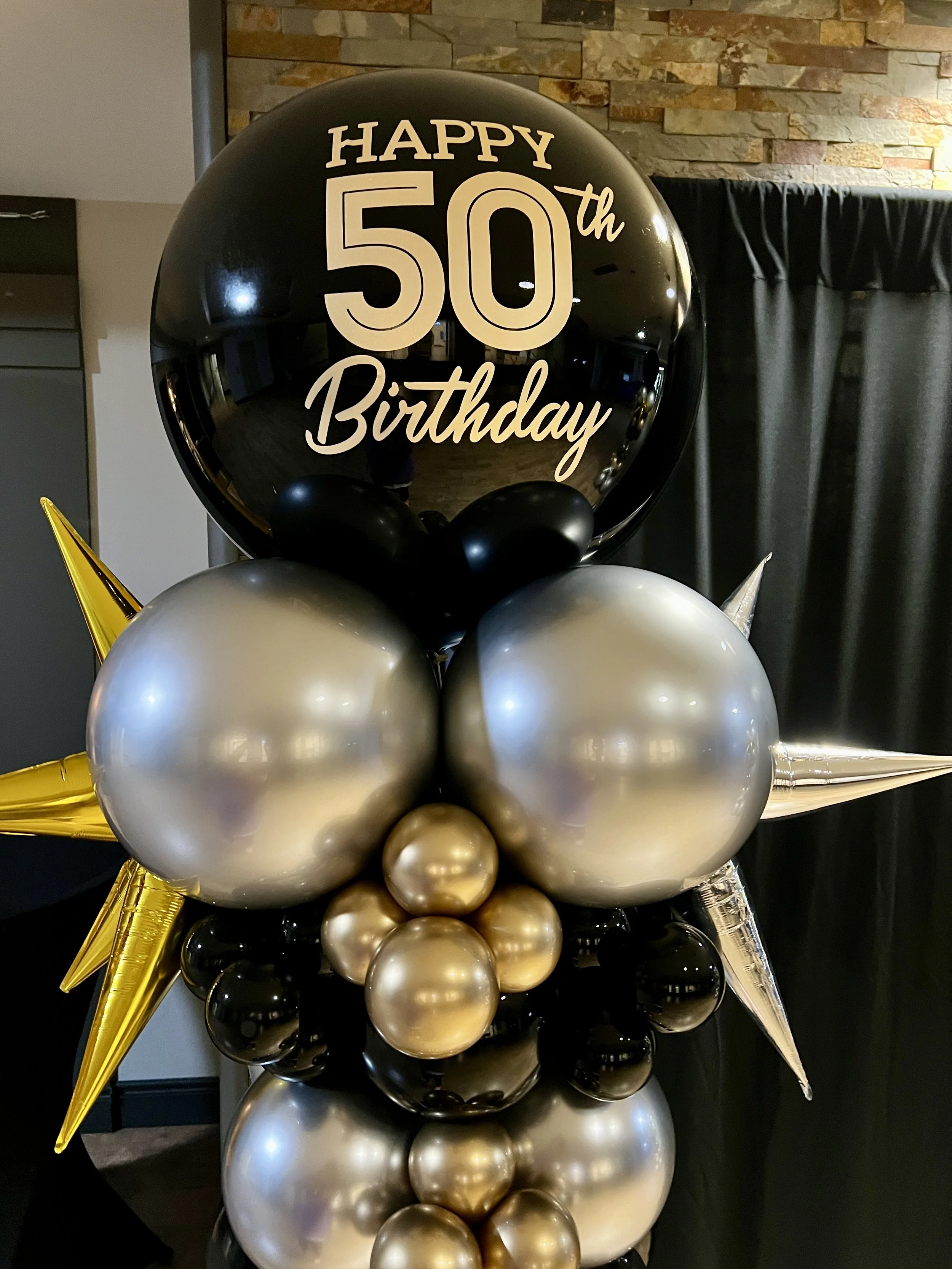 Balloon arrangement for a 50th birthday celebration with black, gold, and silver balloons, including a large black balloon with 'Happy 50th Birthday' custom vinyl. 