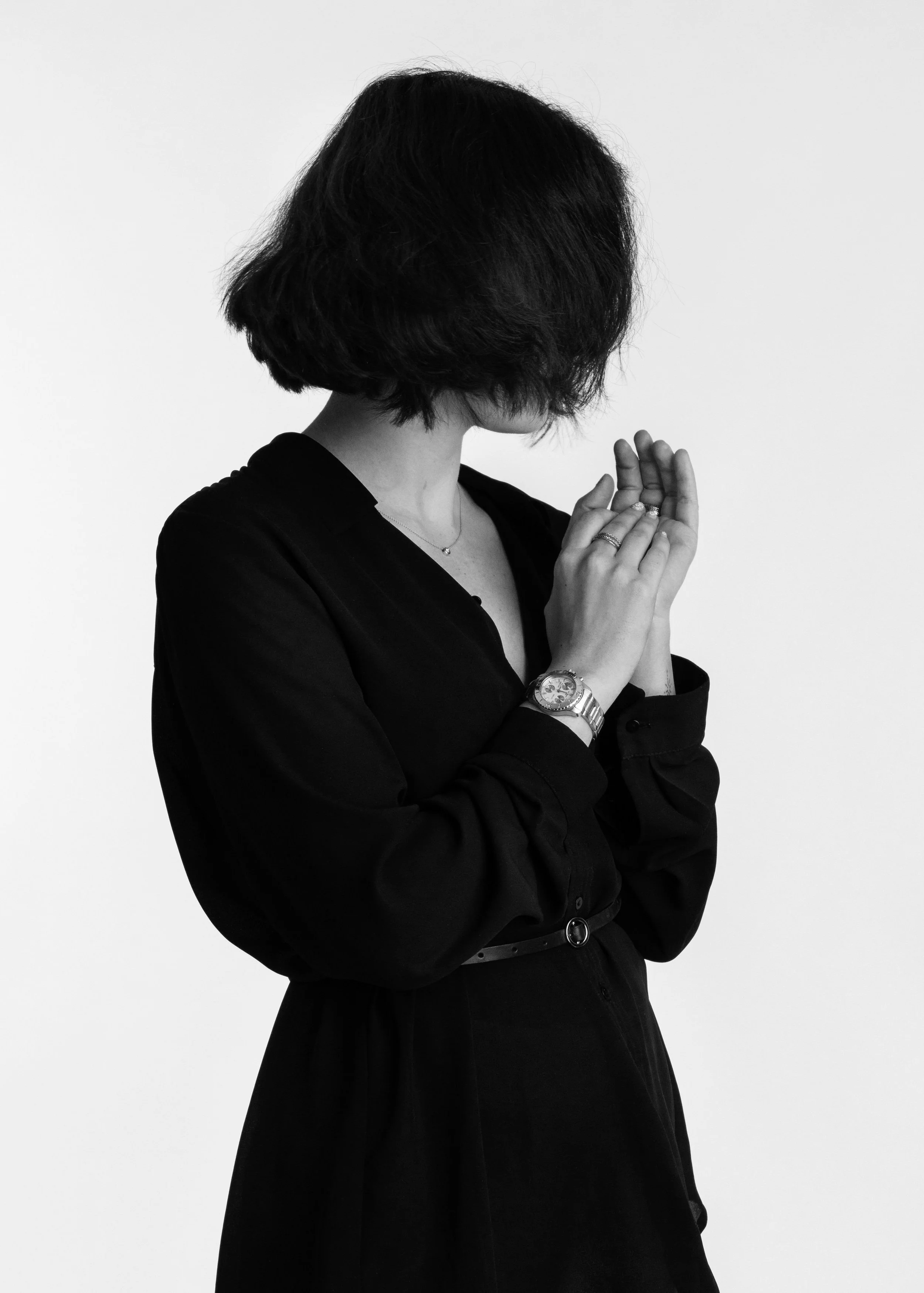 Black and white portrait of a woman with a bob haircut, face turned down and hidden, hands clasped near her chin, wearing a black dress and a wristwatch