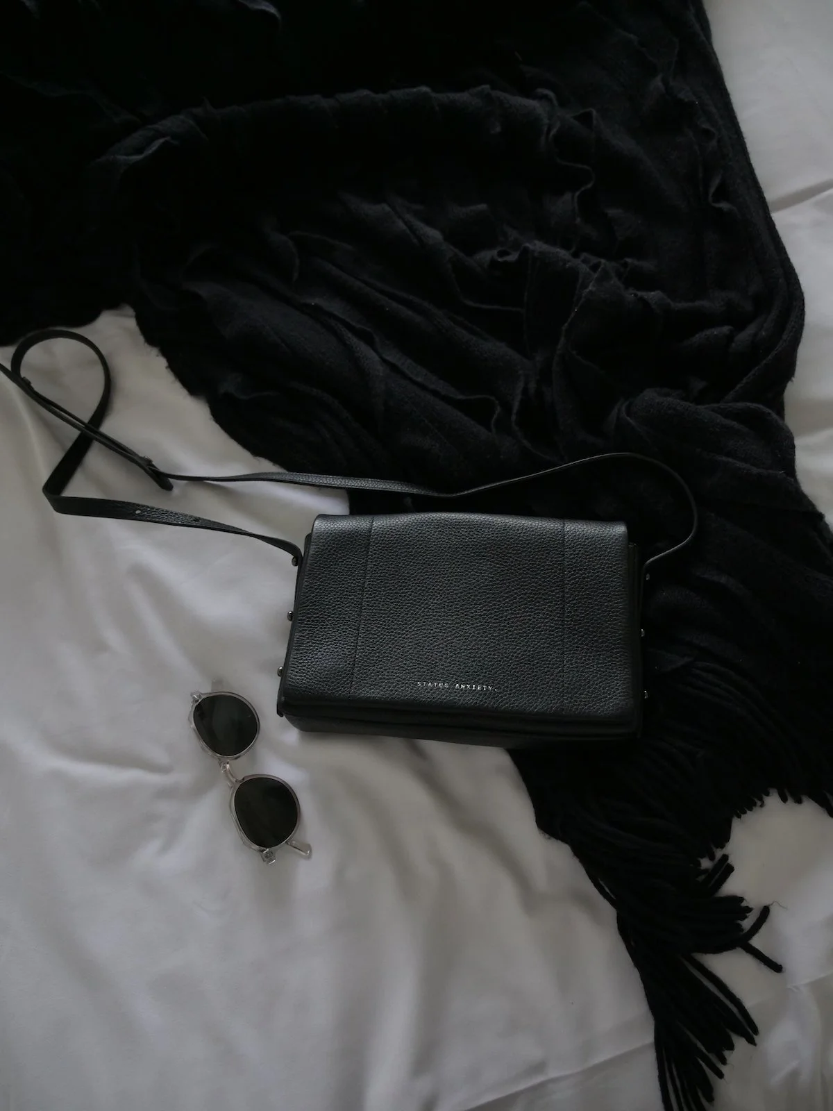 Flat-lay photo of a black crossbody bag, sunglasses, and a black fringed wrap on a white bed