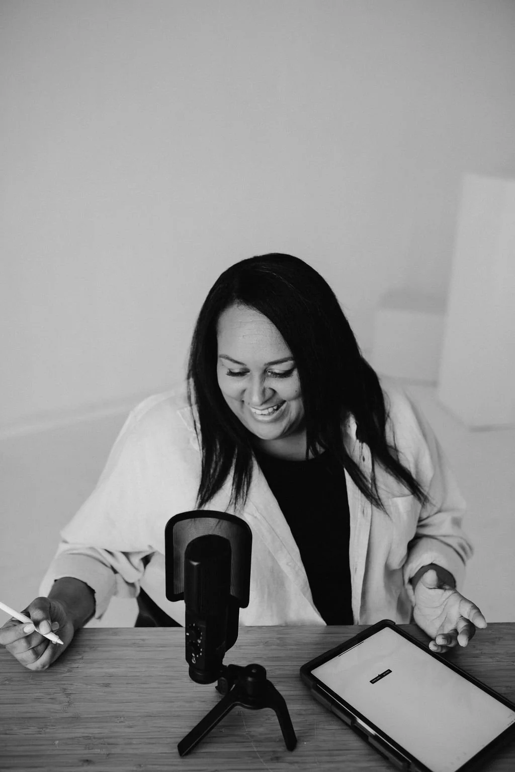 Black and white photo of Sherise Adkins smiling at her desk with a podcast microphone and tablet, holding a pen