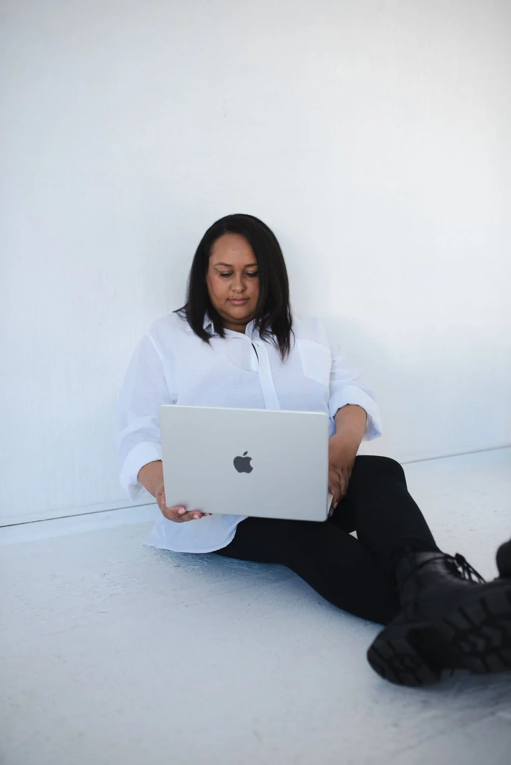 Sherise Adkins sitting on the floor against a plain white wall, wearing a white blouse and black trousers, working on a laptop