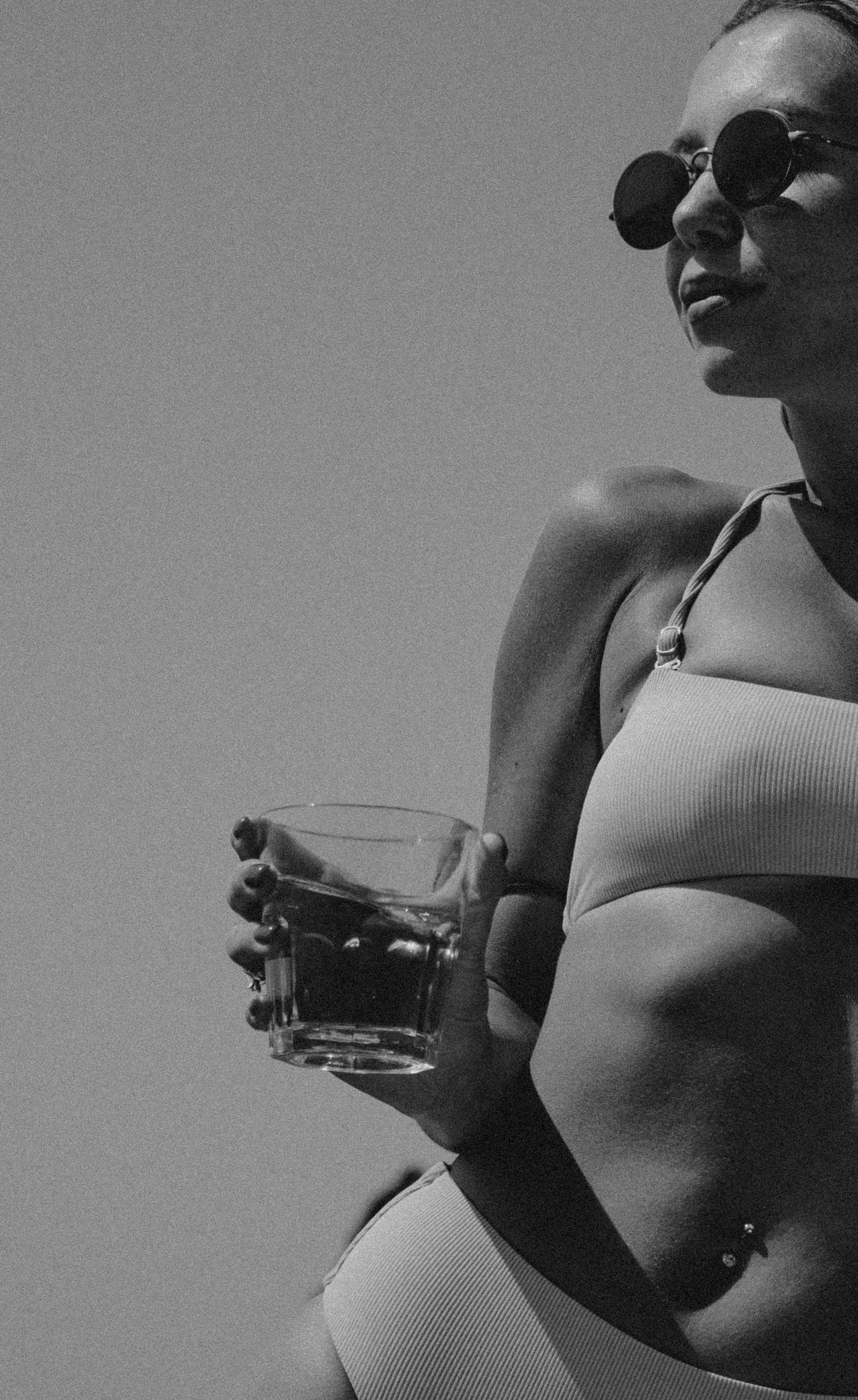 Black and white close-up photo of a woman's torso and shoulder, wearing a ribbed white top with piercings visible, holding a glass jar