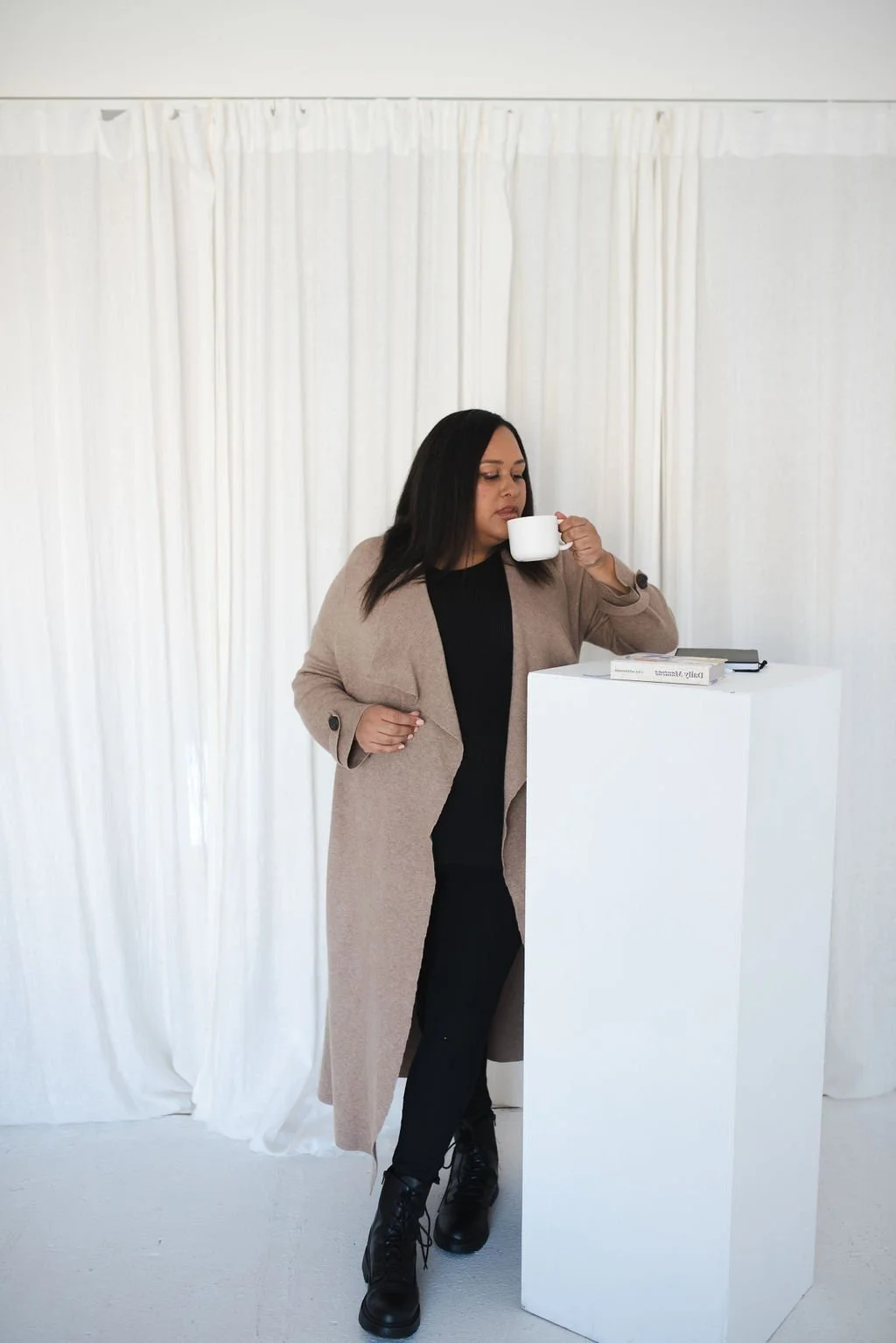Sherise Adkins standing beside a white pedestal with a book, holding a coffee cup, wearing a camel cardigan