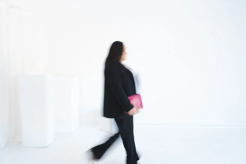 Motion-blur photo of Sherise Adkins walking through a white studio in a black blazer and trousers, holding a pink clutch