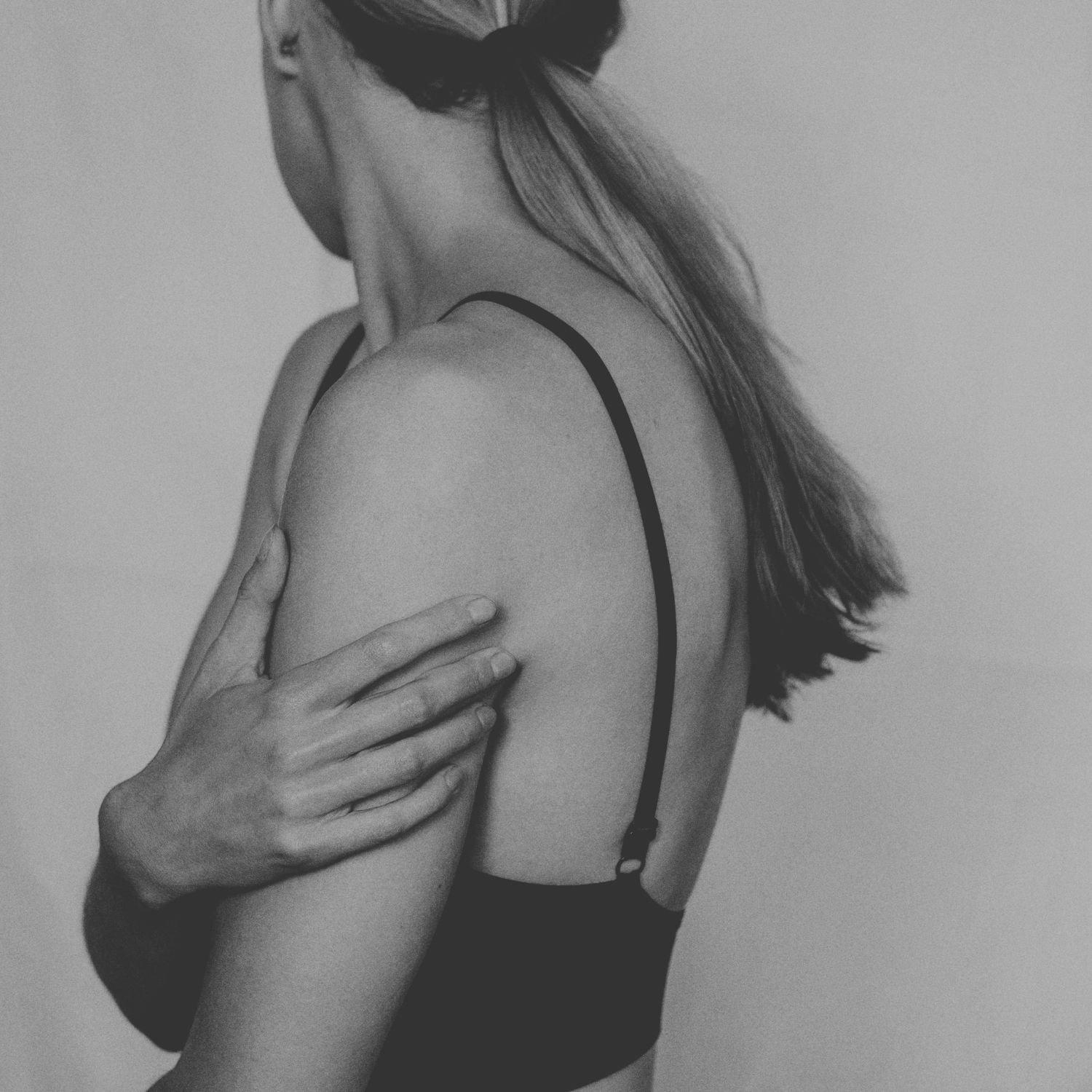 Black and white photo of a woman seen from behind in a black sports bra, blonde hair in a ponytail, one hand resting on her opposite shoulder, against a plain pale backdrop.