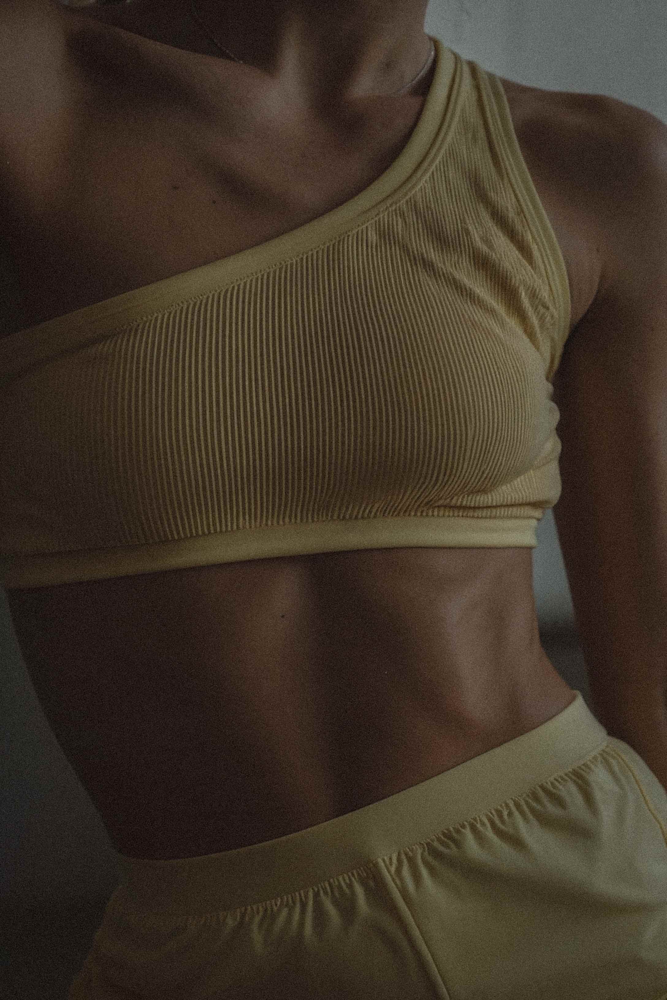 Close-up photo of a woman in a pale yellow ribbed one-shoulder crop top and matching shorts, shot in warm low light against a plain wall.