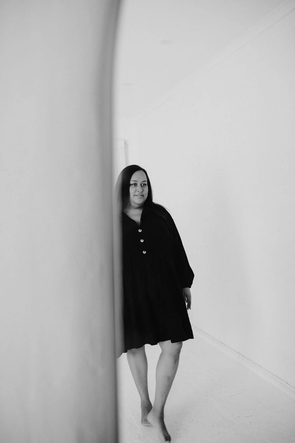 Black and white photo of Sherise Adkins walking barefoot through a bright, curved white hallway, wearing a black button-front dress