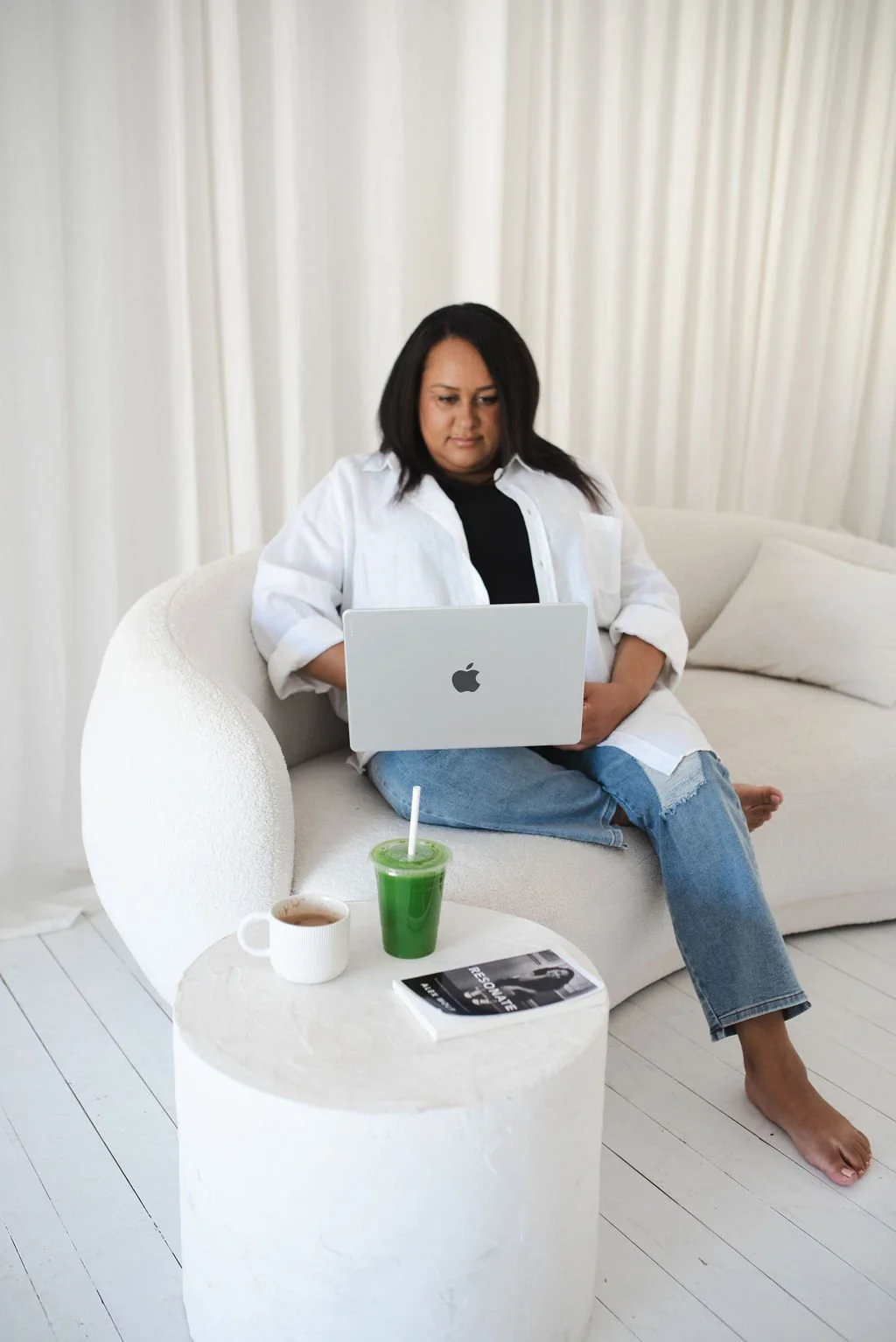 Sherise Adkins sitting on a curved white couch working on a MacBook, barefoot in jeans and a white shirt, with a coffee, green juice and book on a round white side table in front of a white curtain backdrop