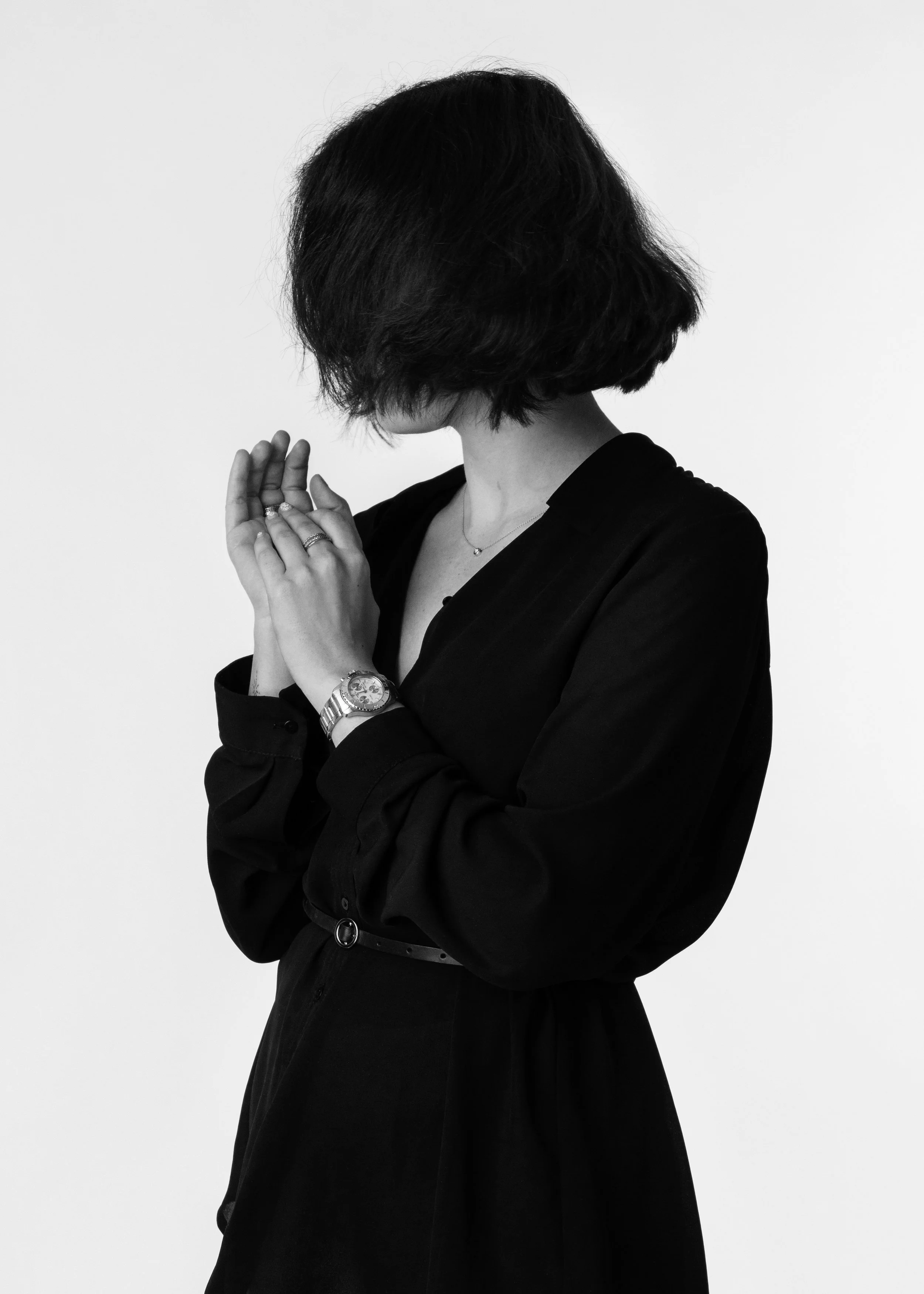 Black and white photo of a woman in a black dress with a bob haircut, face turned away, hands clasped together and wearing a wristwatch