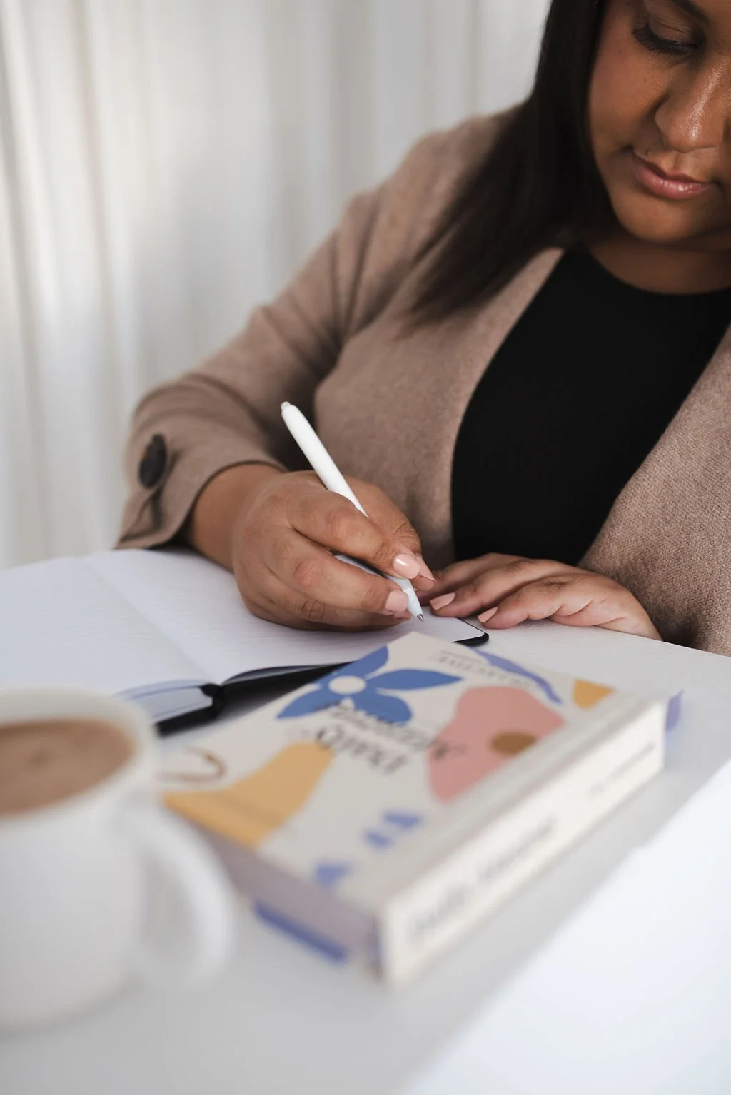 Photo of a woman in a tan blazer writing in a notebook at a table with a coffee cup and a book