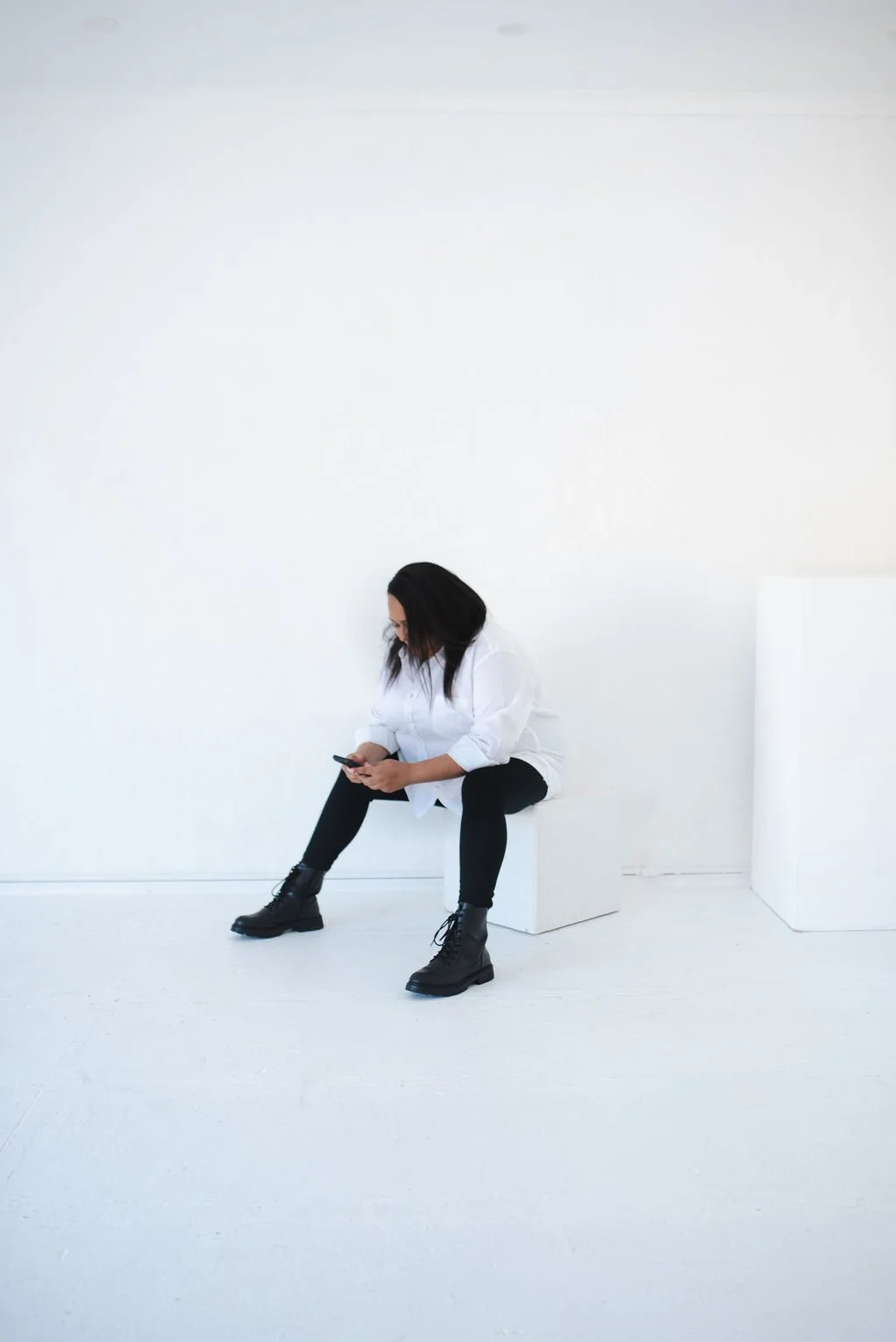 Sherise Adkins sitting on a white cube in a minimalist white studio, looking down at her phone, wearing a white shirt and black boots