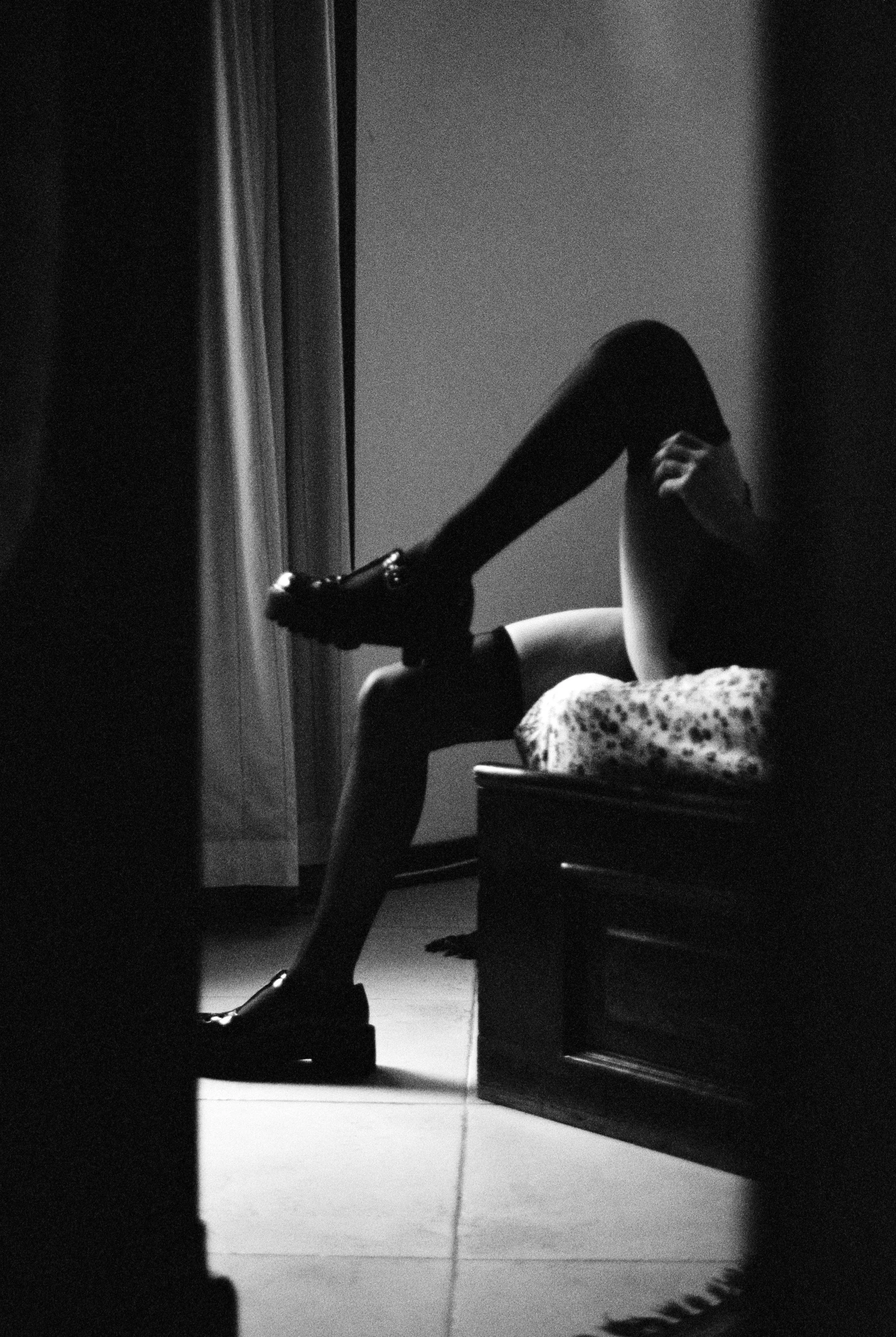 Black and white photo of a woman's legs, one shoe on and one shoe on the floor, seated on the edge of a bed near a curtained window