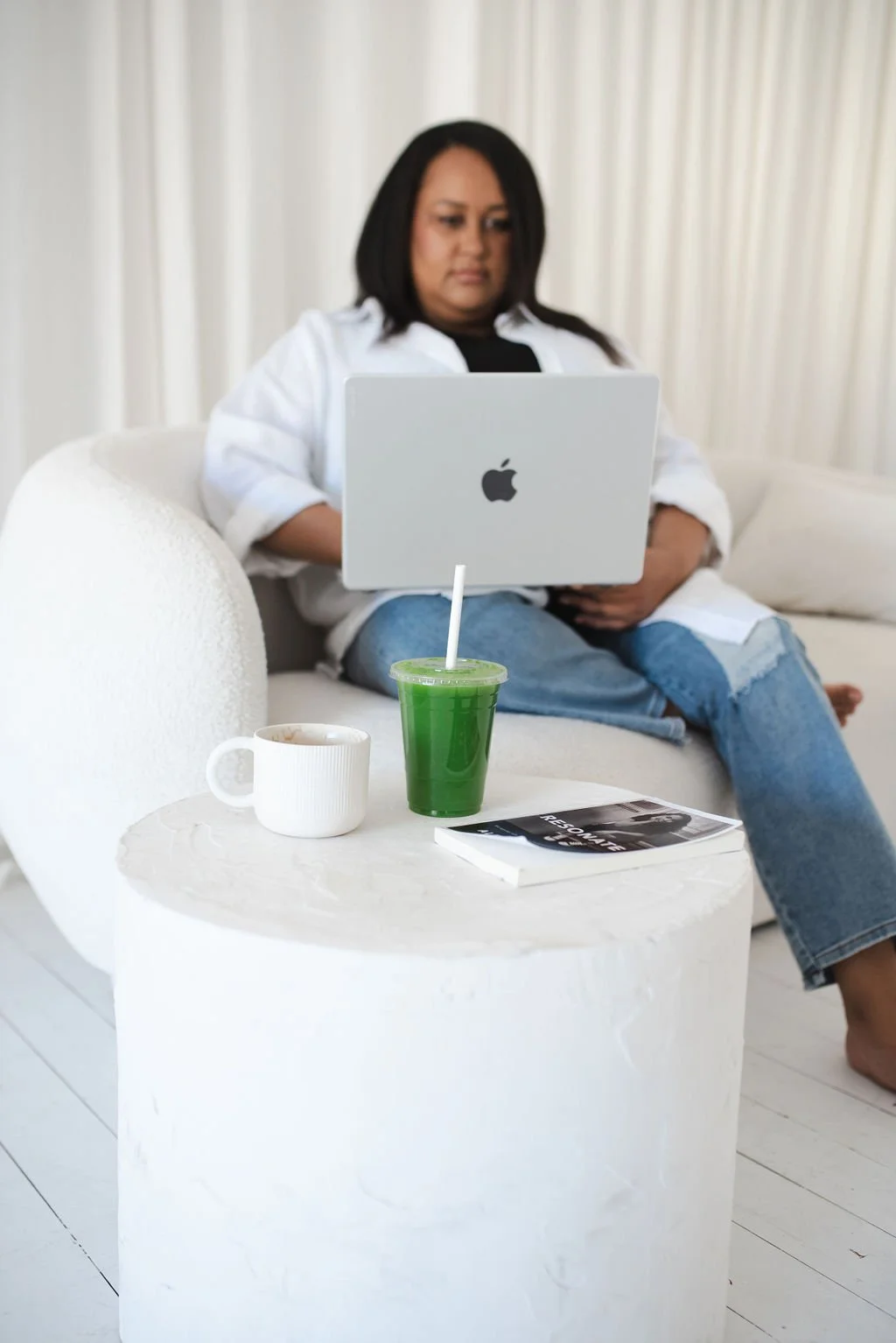 Sherise Adkins sitting on a cream sofa working on a laptop, with a green juice, coffee and magazine on a round side table beside her