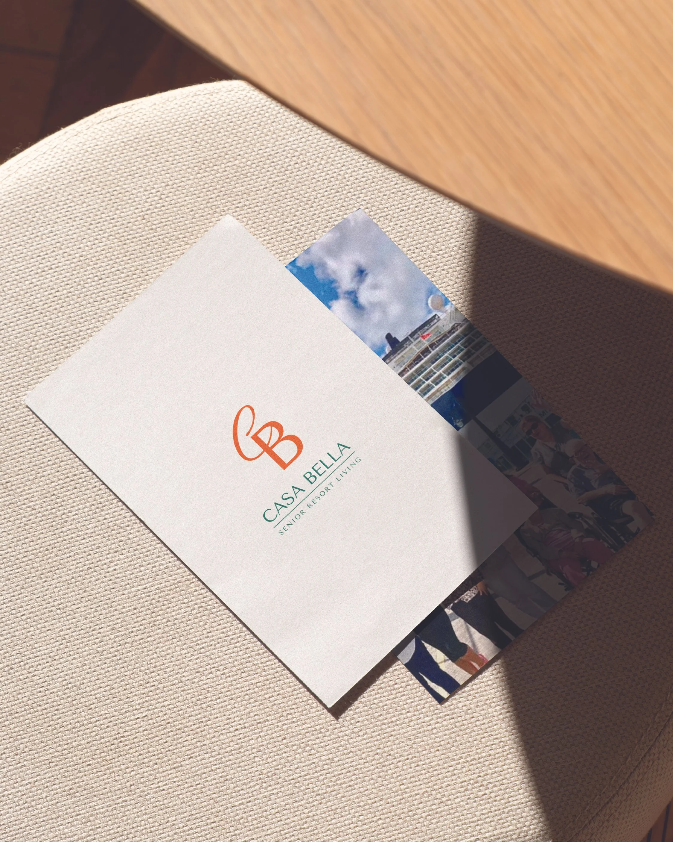 A brochure for Casa Bella Senior Resort Living, placed on a light-colored cushion with a wooden surface above it. The brochure has a logo with the letters 'CB' and the text 'Casa Bella Senior Resort Living'. Behind the brochure, there are some photos