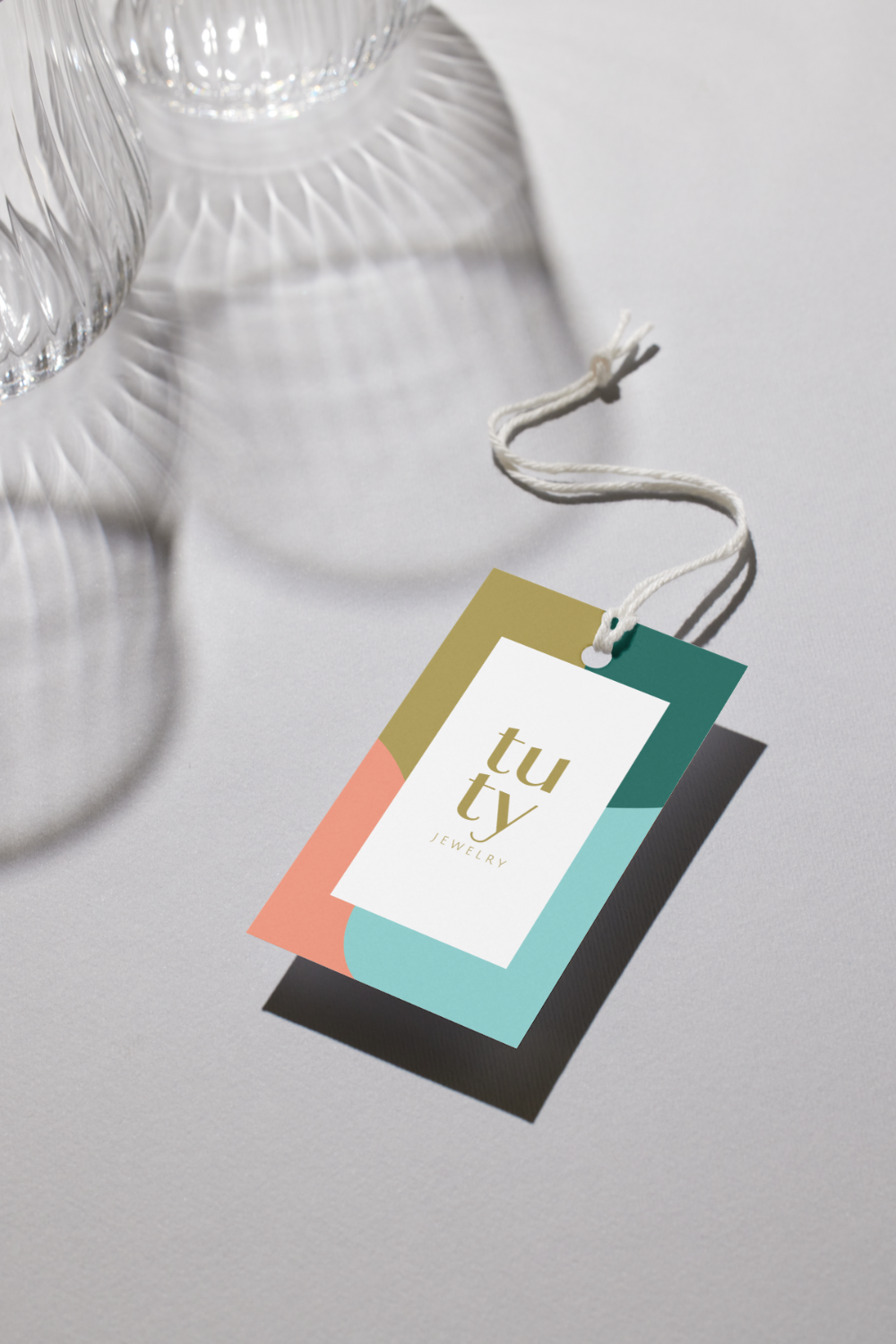 A colorful jewelry tag with the brand name 'tu tv' and the word 'JEWELRY' underneath, lying on a white surface with a shadow. Part of a glass object is visible in the upper left corner.
