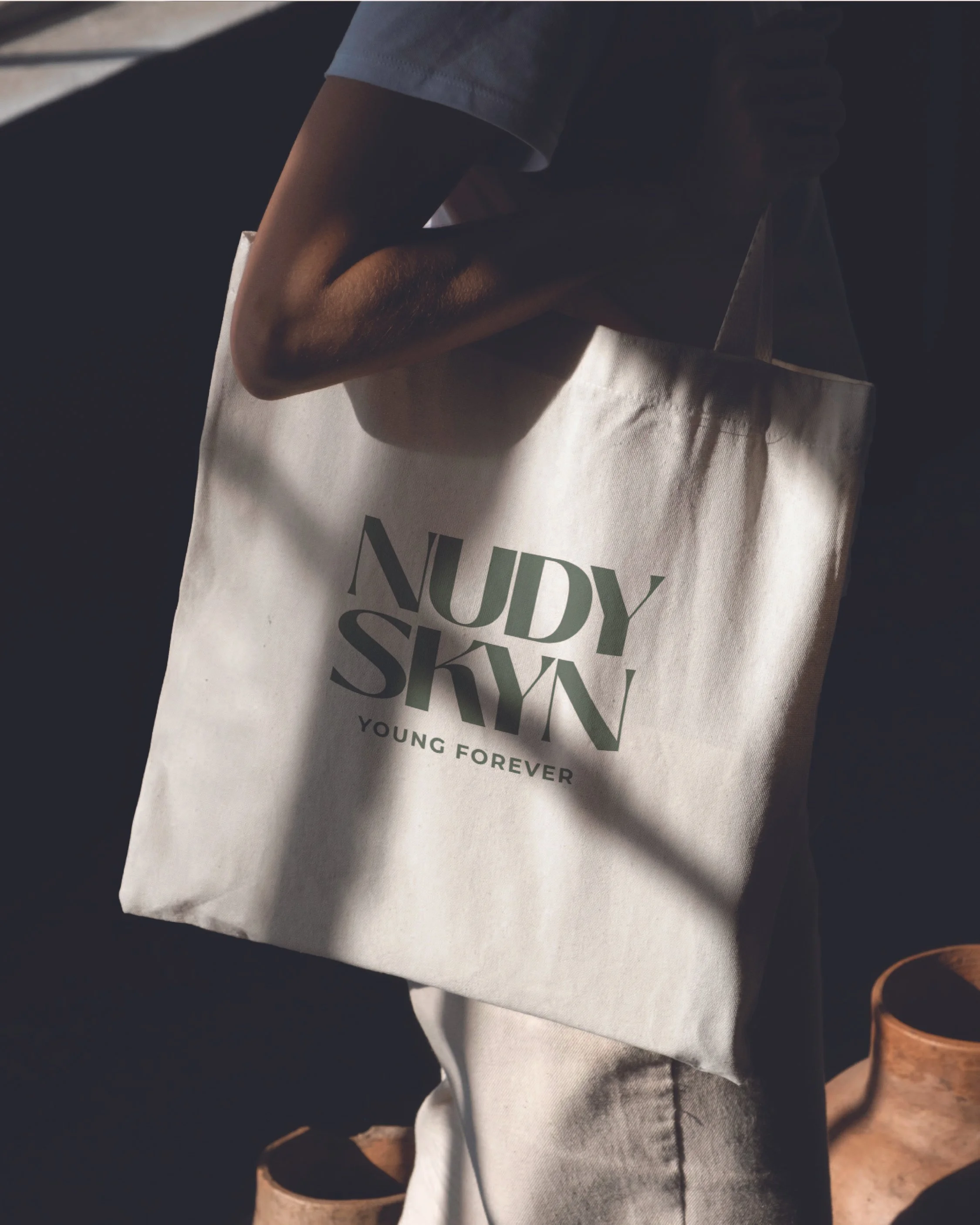 Person holding a white canvas tote bag with the text 'Nudy Skin' and 'Young Forever' printed on it, in a dimly lit environment.