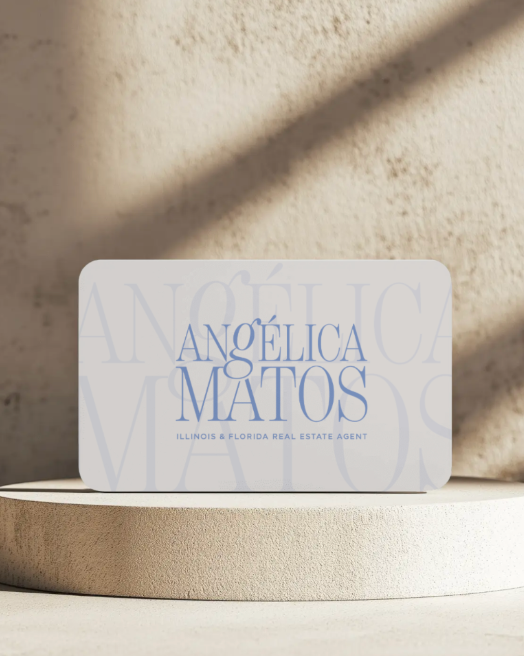 Business card for Angélica Matos, Illinois & Florida real estate agent, placed on a beige surface with a textured background and shadow.