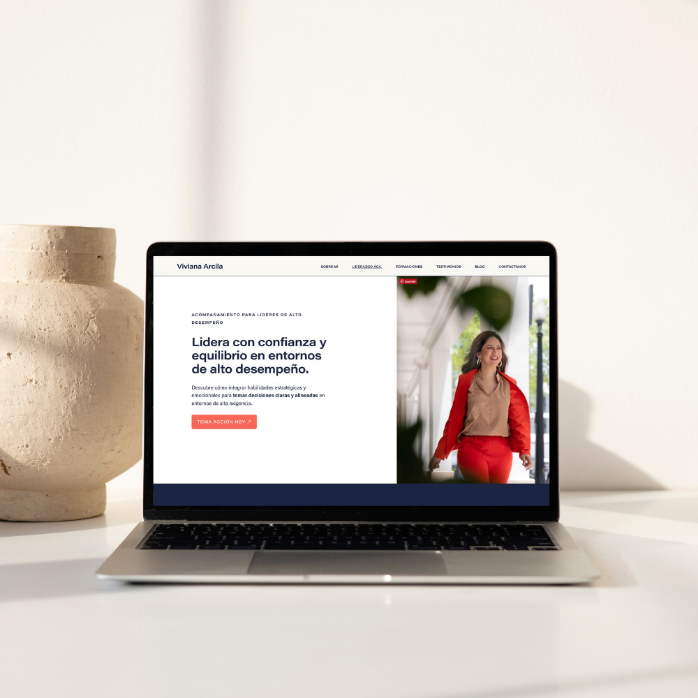 Laptop on a white table displaying a website about leadership coaching, with a woman walking outdoors on the screen, beside a large beige vase.