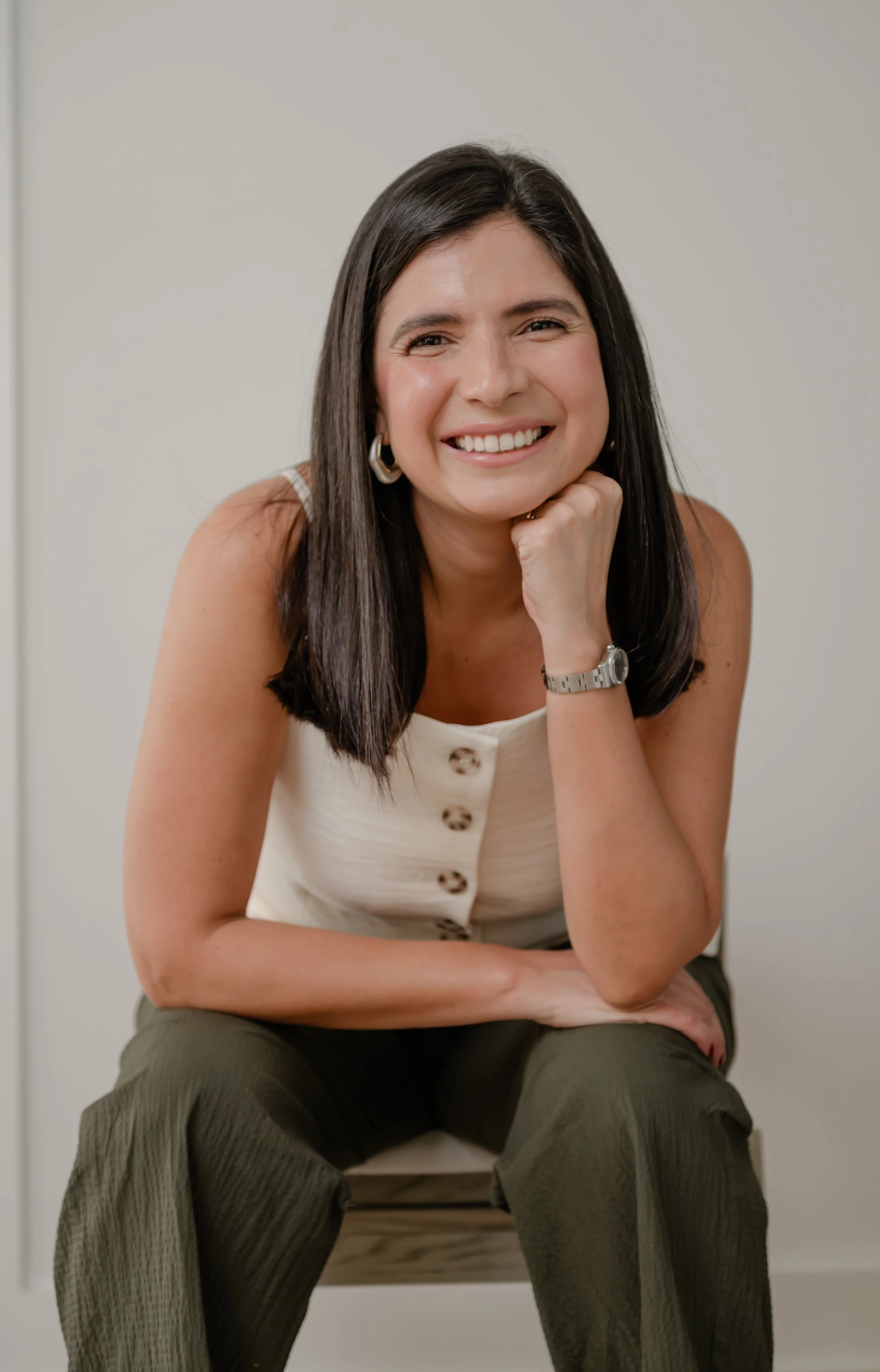 Portrait of a woman with dark hair smiling, sitting on a chair with her chin resting on her hand, wearing a sleeveless top and dark pants.