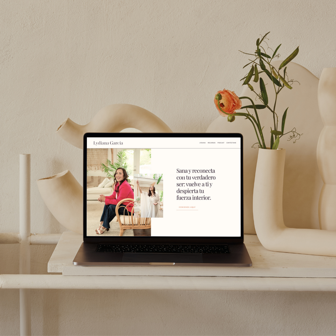 Laptop on a white desk displaying a website with a woman in a pink jacket and white background with plants. Decor includes beige vase with pink flower and abstract sculpture on a cream wall.