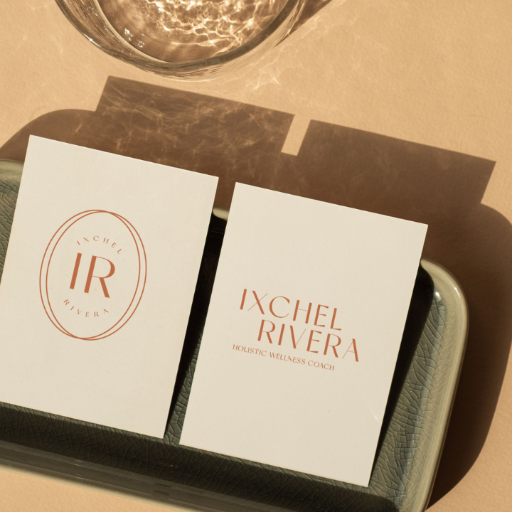 Two white cards on a tray, one with a logo reading 'IR' and 'IXCHEL RIVERA', the other with the text 'IXCHEL RIVERA HOLISTIC WELLNESS COACH'. A glass of water is above and to the left, casting shadows on the cards.