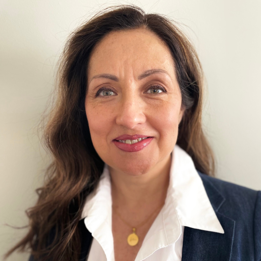 A woman with long, wavy brown hair, light skin, and hazel eyes, wearing a white collared shirt and a dark blazer, smiling at the camera against a plain, light background.