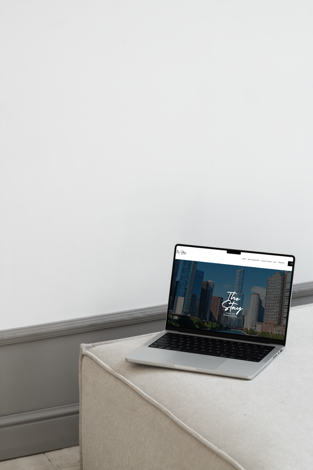 A silver laptop with a black keyboard displaying a website with a cityscape and the text 'The Stay' on a beige fabric-covered surface in a minimalistic room.