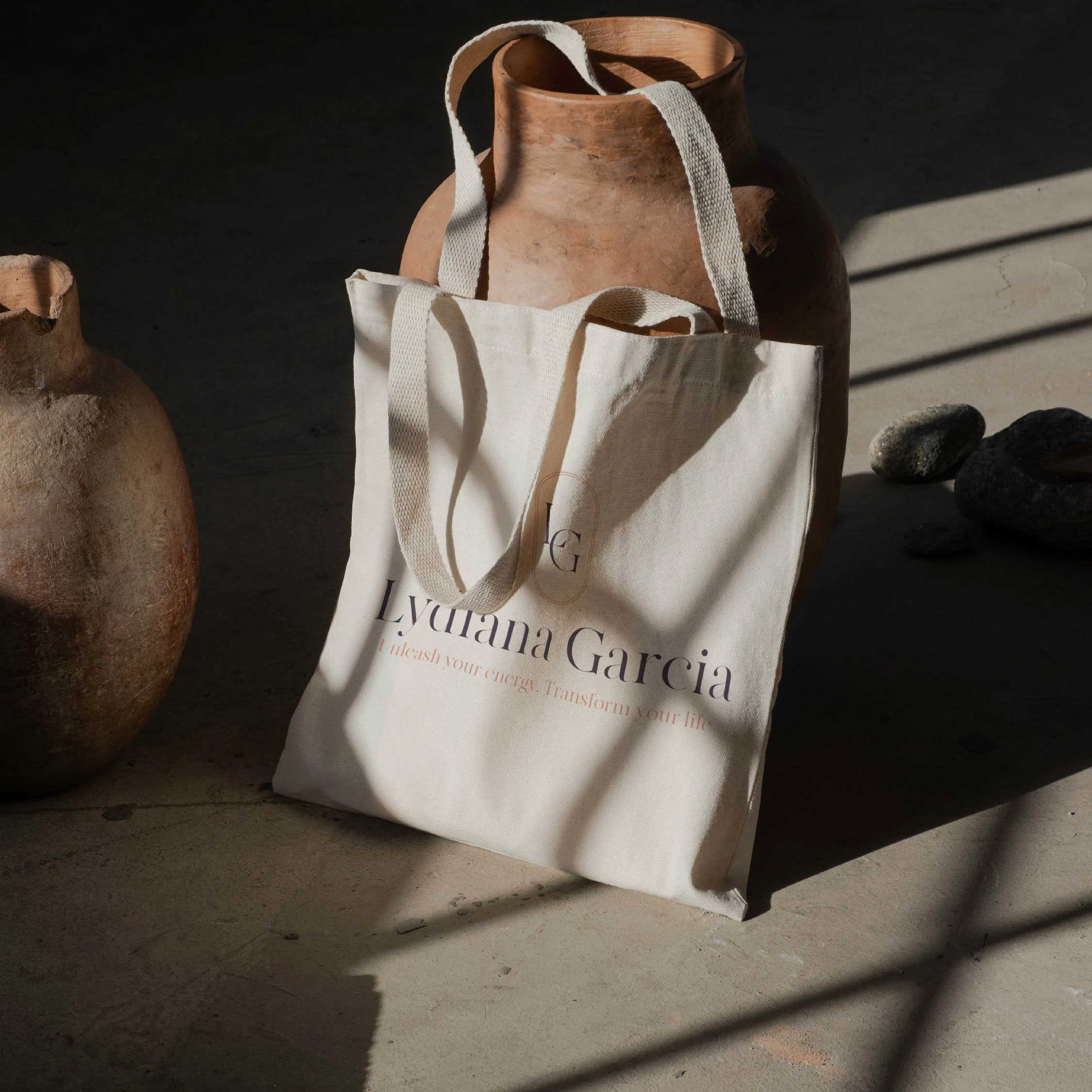 A tote bag with the name 'Luyana Garcia' printed on it, placed on a concrete surface with sunlight and shadows. There are two large, rustic clay vases and some stones nearby.