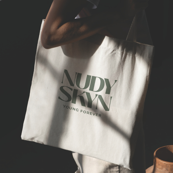 Person holding a white tote bag with the words "NUDY SKIN" and "YOUNG FOREVER" printed on it, with a shadow cast across the bag.