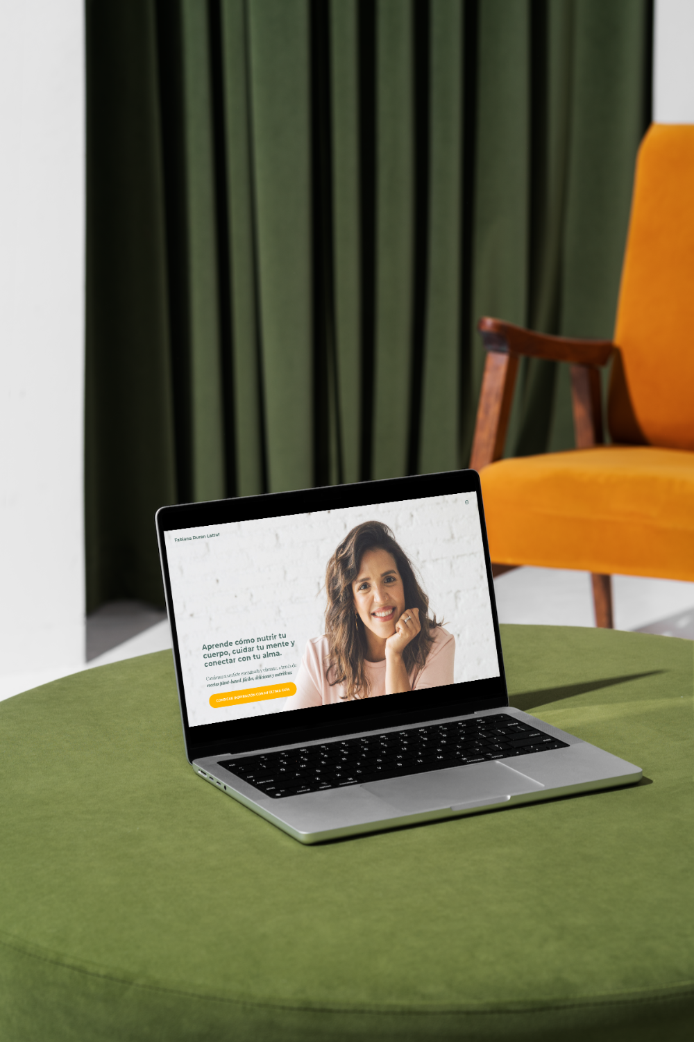 Laptop on a green table displaying a woman and Spanish text about nutrition and mental health, with a yellow call-to-action button. Background includes a yellow chair and green curtains.