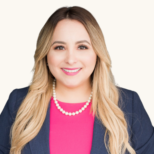 A professional woman with blonde hair, wearing a navy blazer, pink top, and pearl necklace, smiling against a white background.