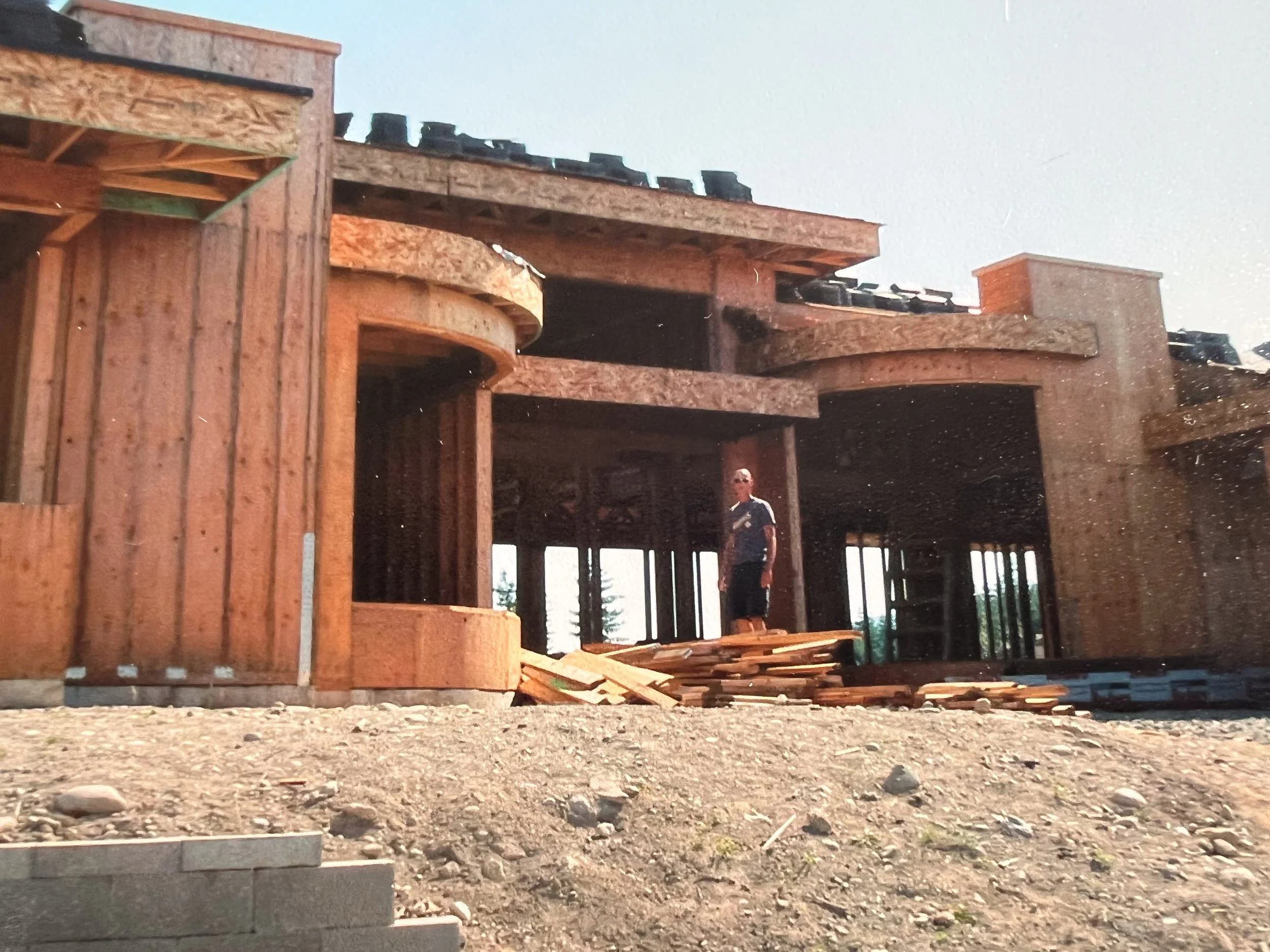 A man standing in front of a house under construction, with wooden framework and plywood on the roof, on a dirt lot with scattered wood pieces.
