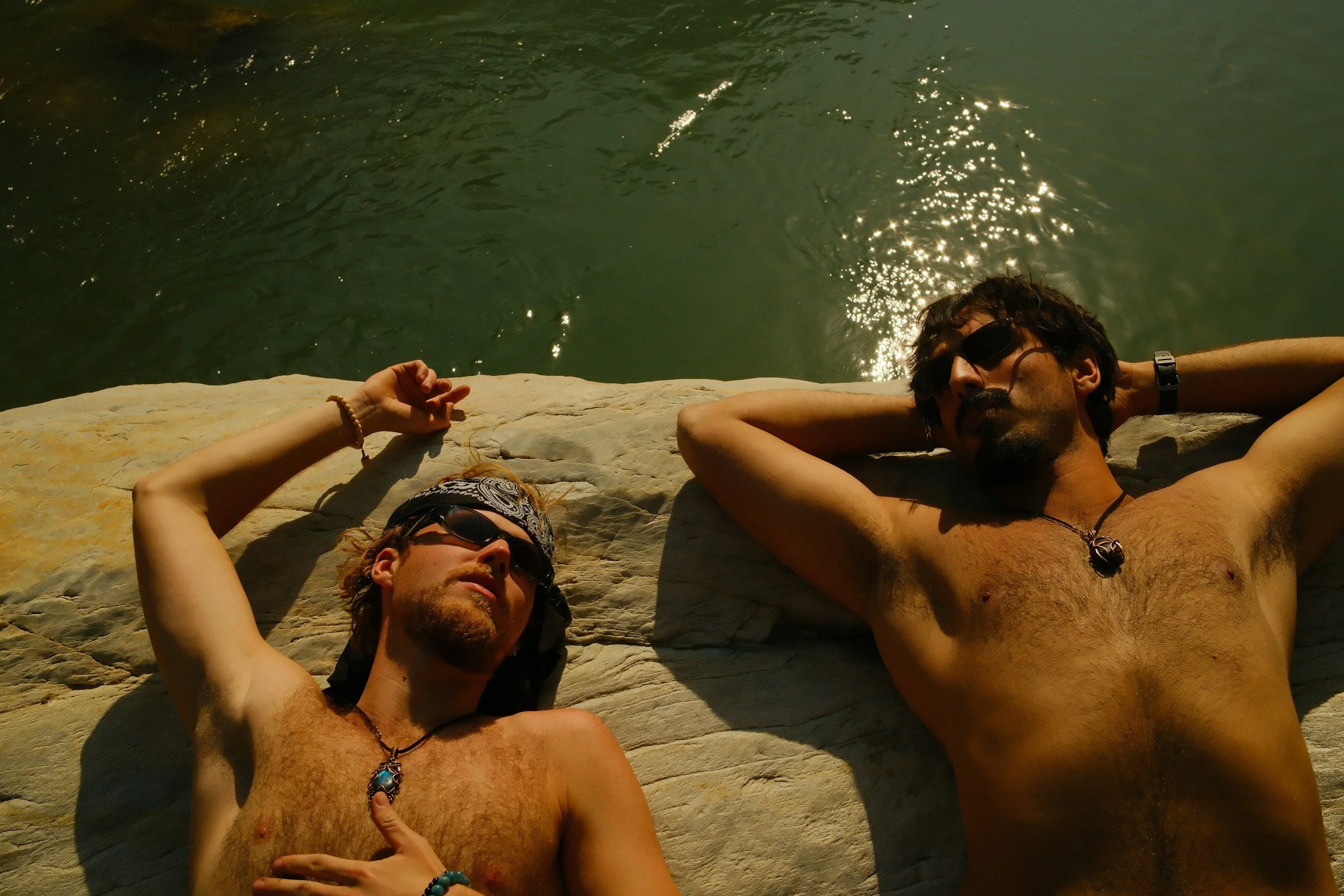 Two shirtless men lying on a rocky surface near water, wearing sunglasses, with one man touching his chest and the other resting his head on his arm, both relaxed and enjoying the sunny weather.