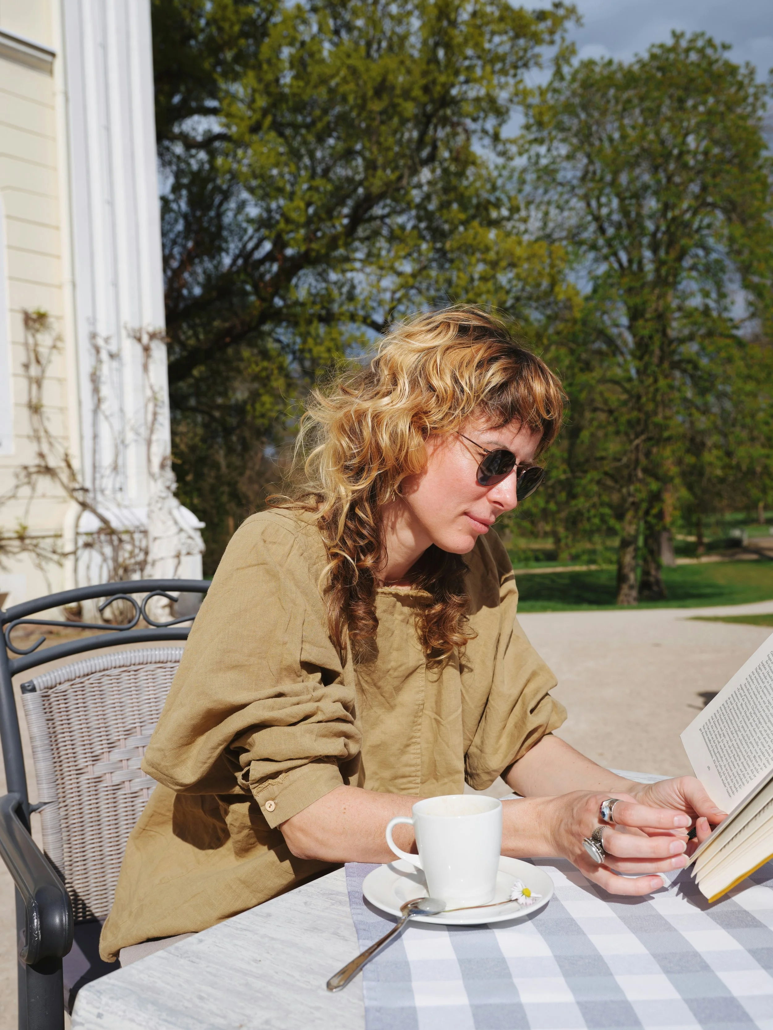 A woman with curly blonde hair and sunglasses sitting at a table outside, reading a book. There is a white mug on a plate with a spoon and a small flower, with a checkered tablecloth. Trees and greenery are visible in the background.