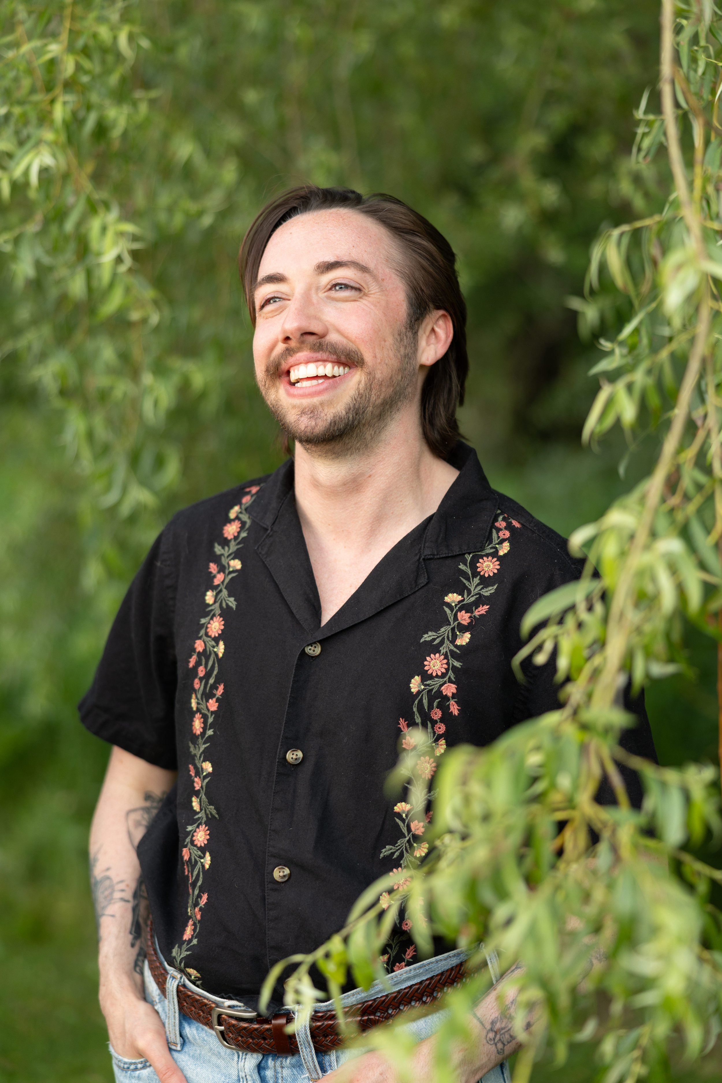 A smiling man with dark hair and facial hair standing outdoors among green foliage, wearing a black shirt with floral embroidery and light blue jeans with a braided belt.
