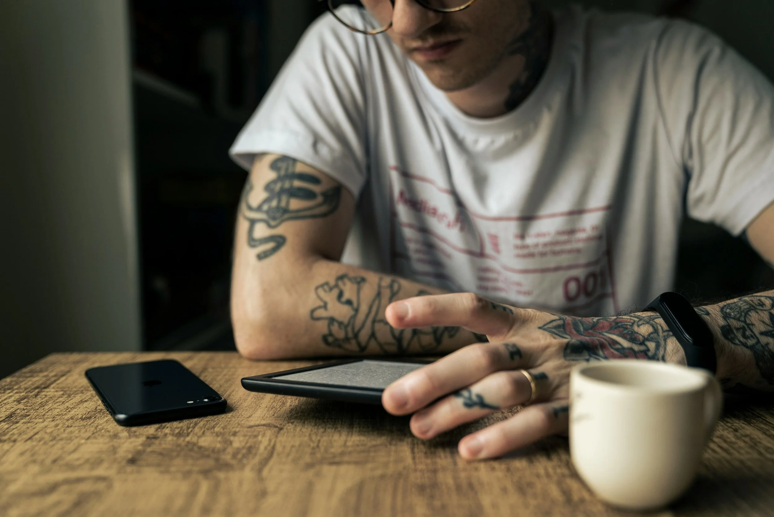 A man with tattoos and glasses is sitting at a wooden table, using a tablet with a smartphone and a coffee cup nearby.