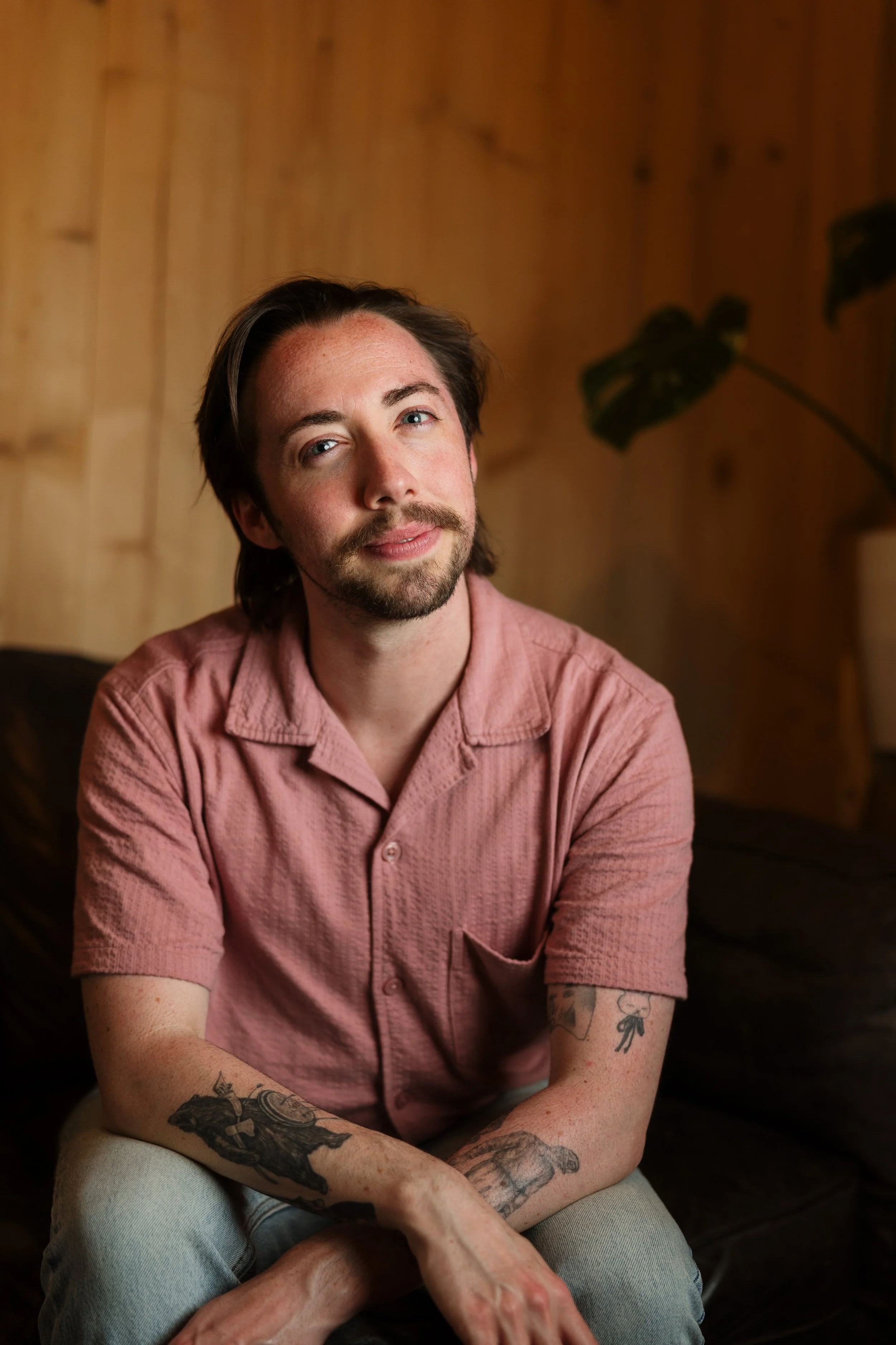 Portrait of a young man with dark hair, beard, and tattoos, wearing a pink shirt and light-colored pants, sitting indoors against a wooden wall with a potted plant in the background.