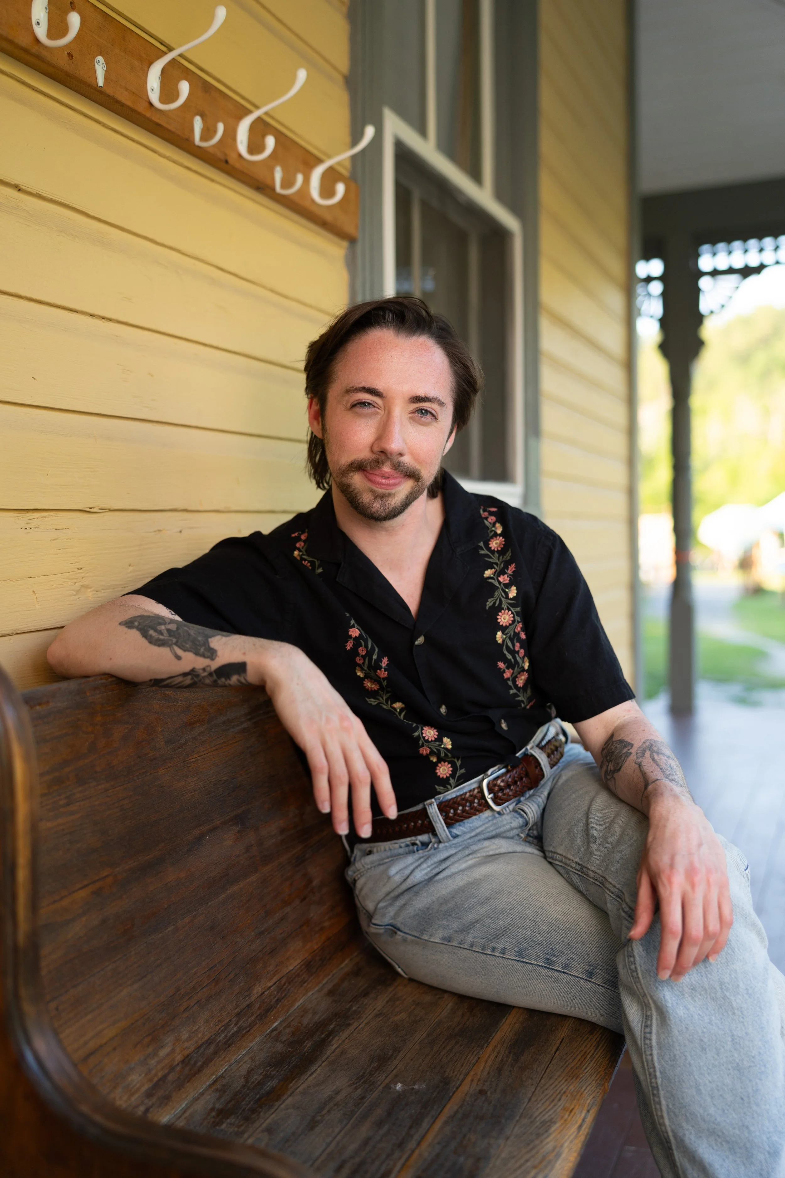 A man with dark hair and tattoos sitting on a wooden bench outside of a yellow house with a porch, smiling and winking at the camera.