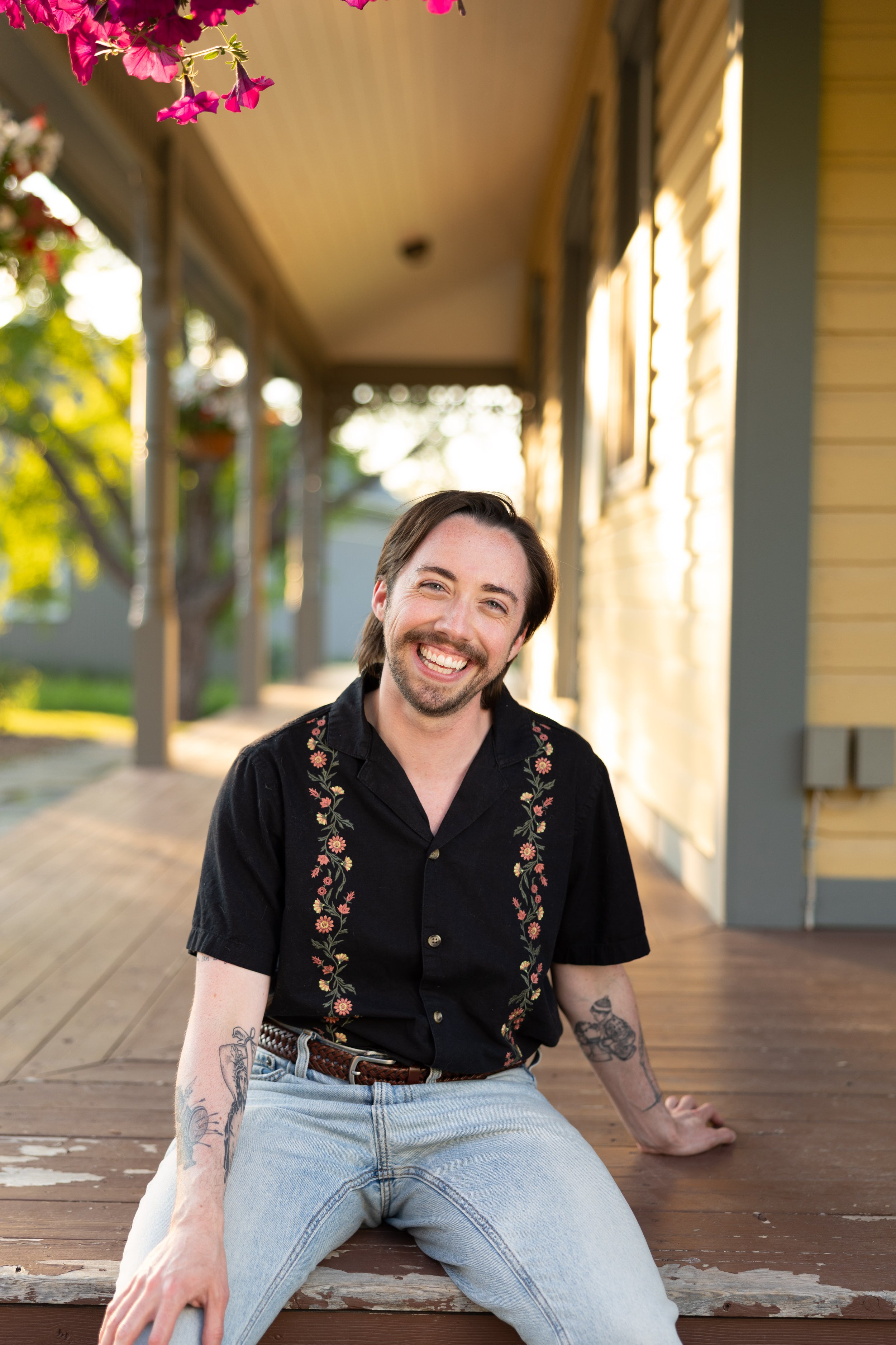 A smiling man with tattoos on his arms, sitting on a wooden porch with a yellow house in the background, during sunset.