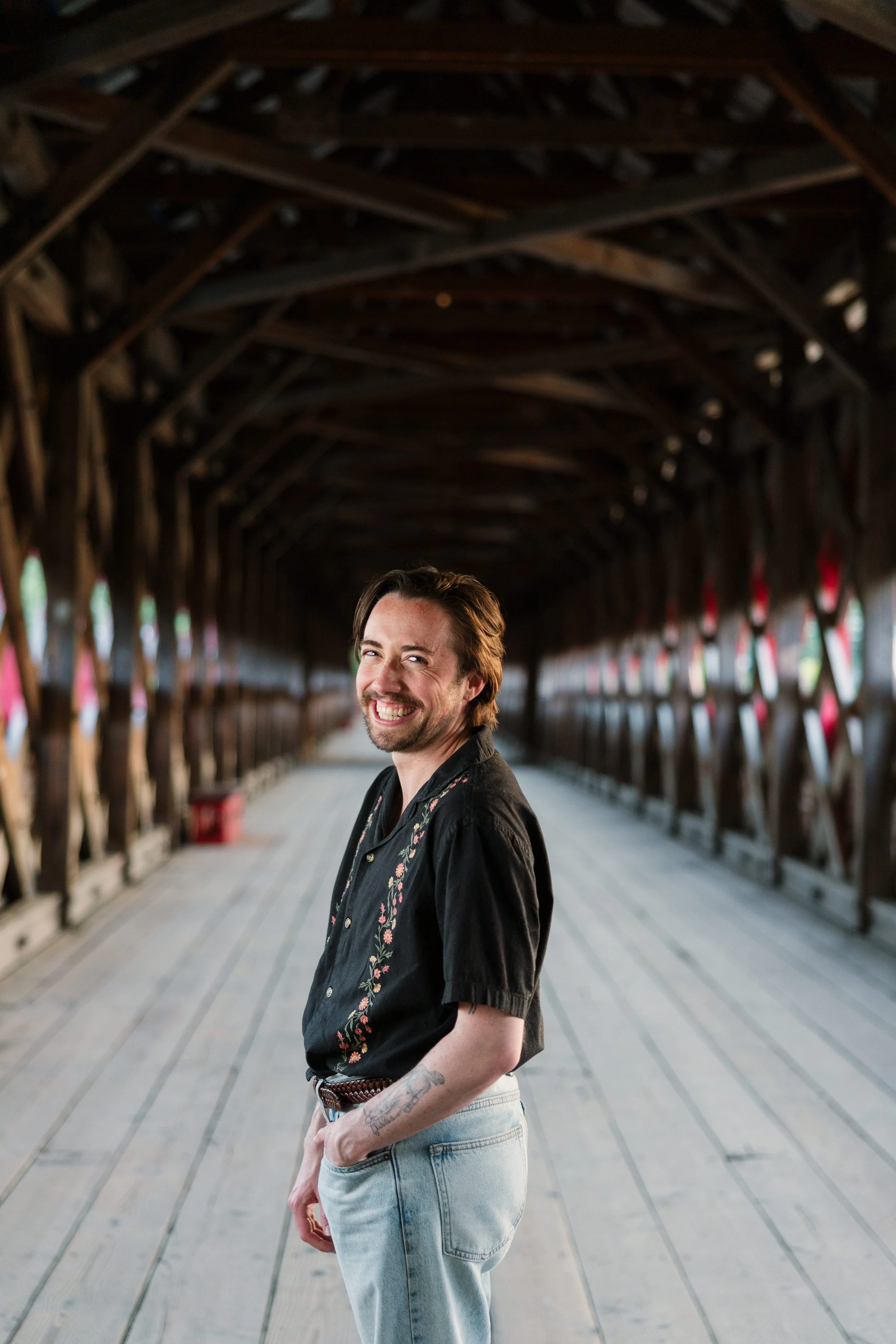 A man with a beard and tattoos on his left arm, smiling and looking at the camera, standing on a covered wooden walkway or bridge with wooden beams overhead.