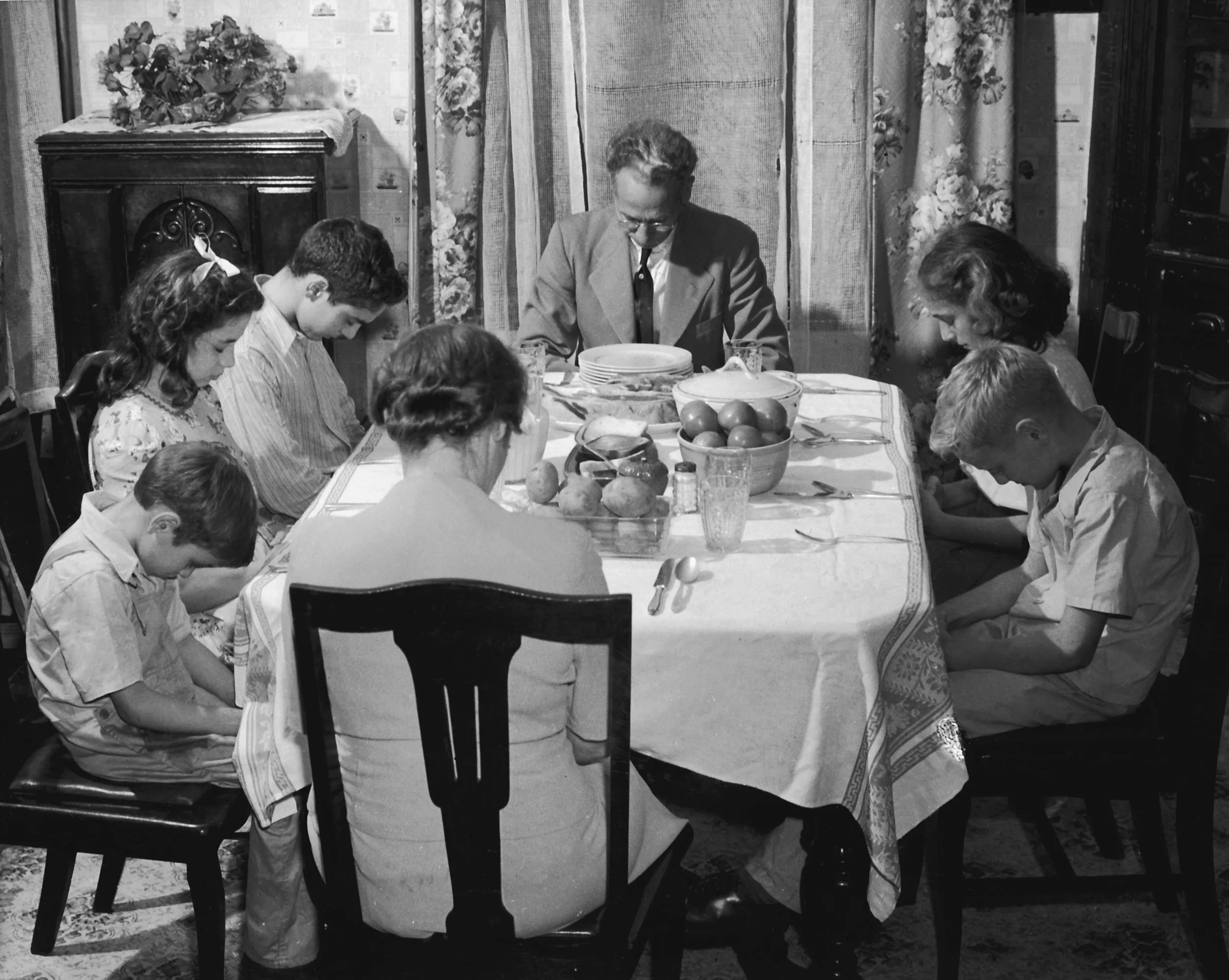 A black and white photo of a family gathered around a dining table during a meal, with a man at the center and various people with bowed heads.