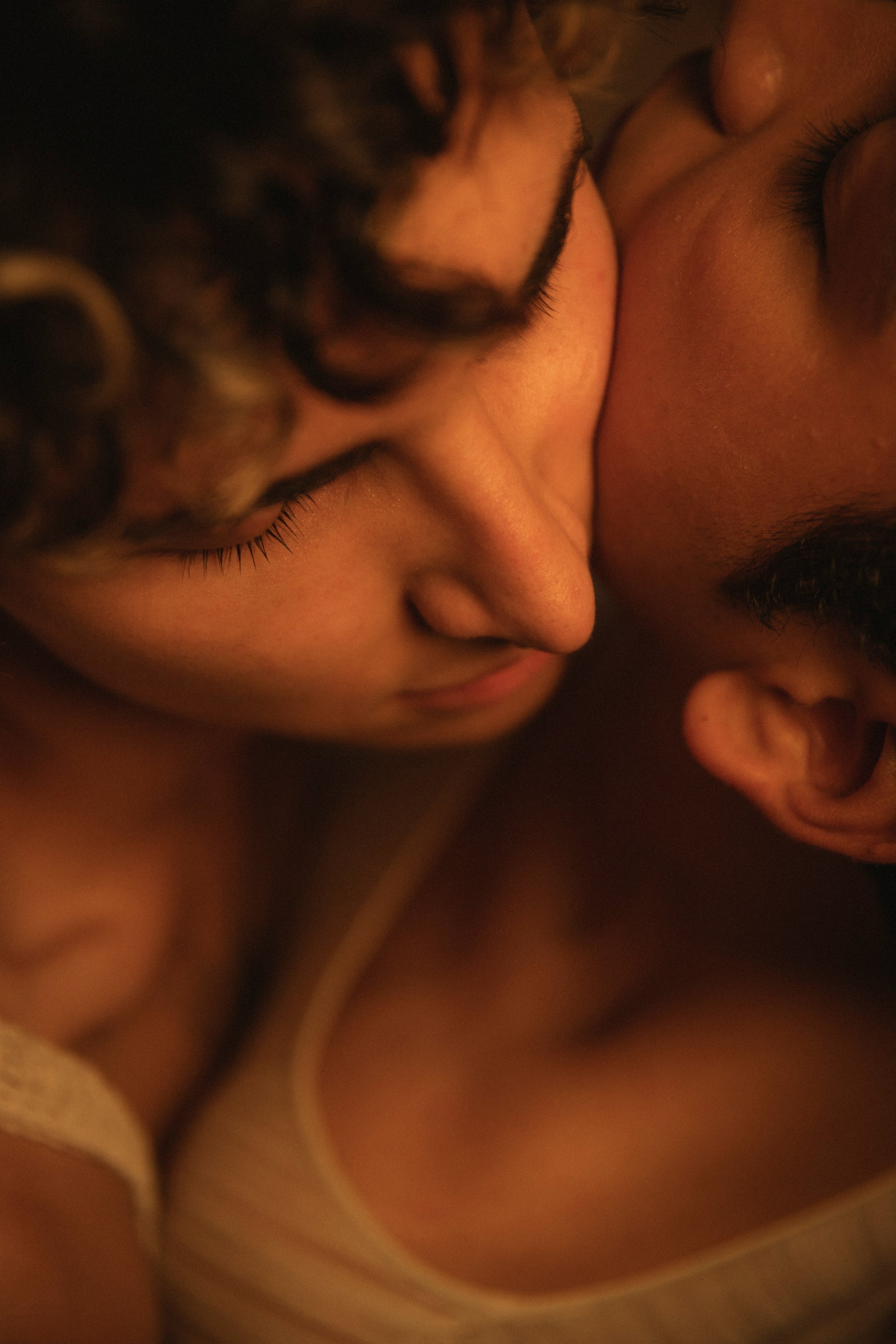 A close-up of two people, one with dark curly hair and the other with dark straight hair, leaning their faces closely together, eyes closed, in an intimate moment.