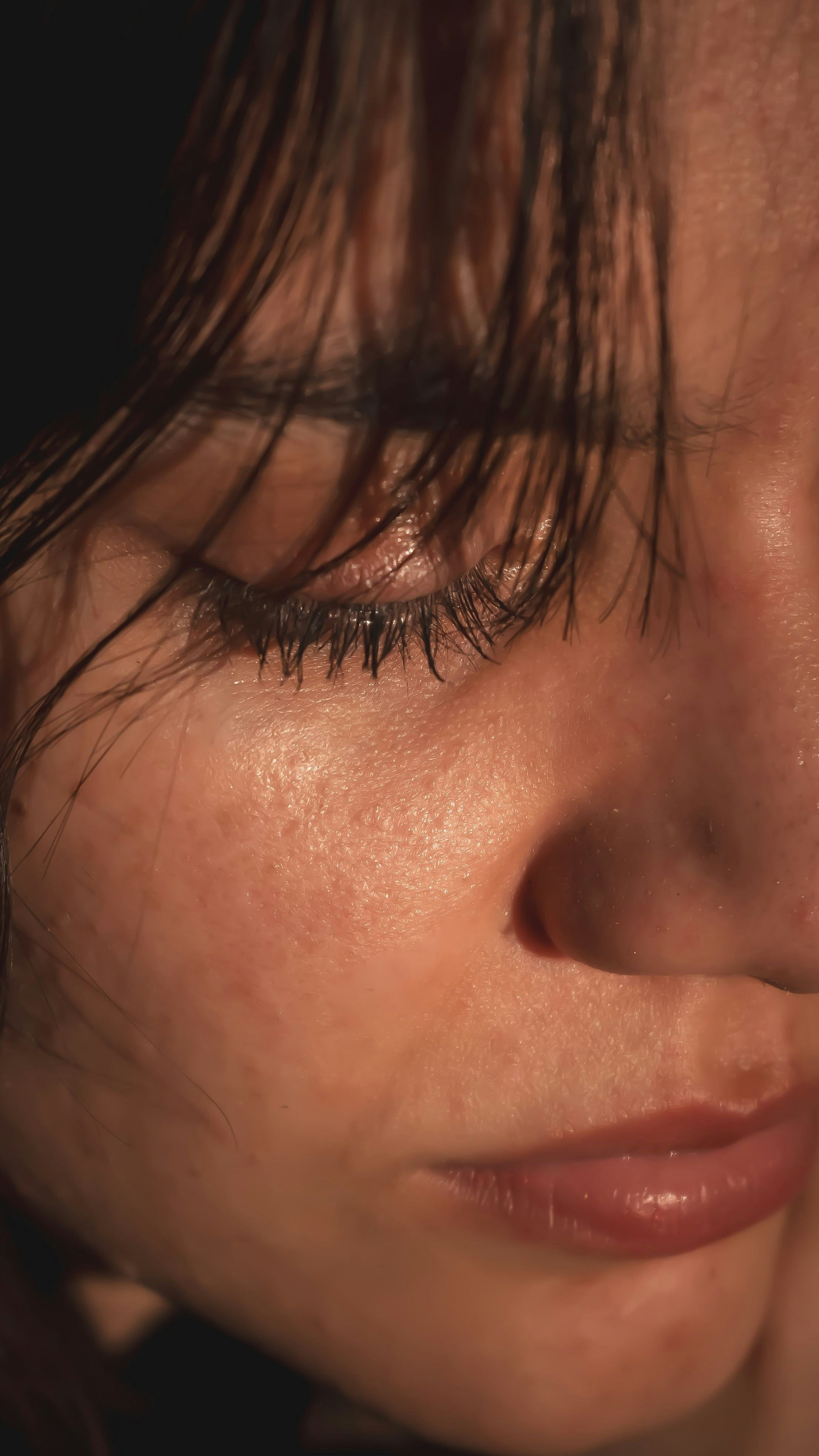Close-up of a person's face with focus on closed eye, wet dark hair covering part of the face, smooth skin, and natural makeup.