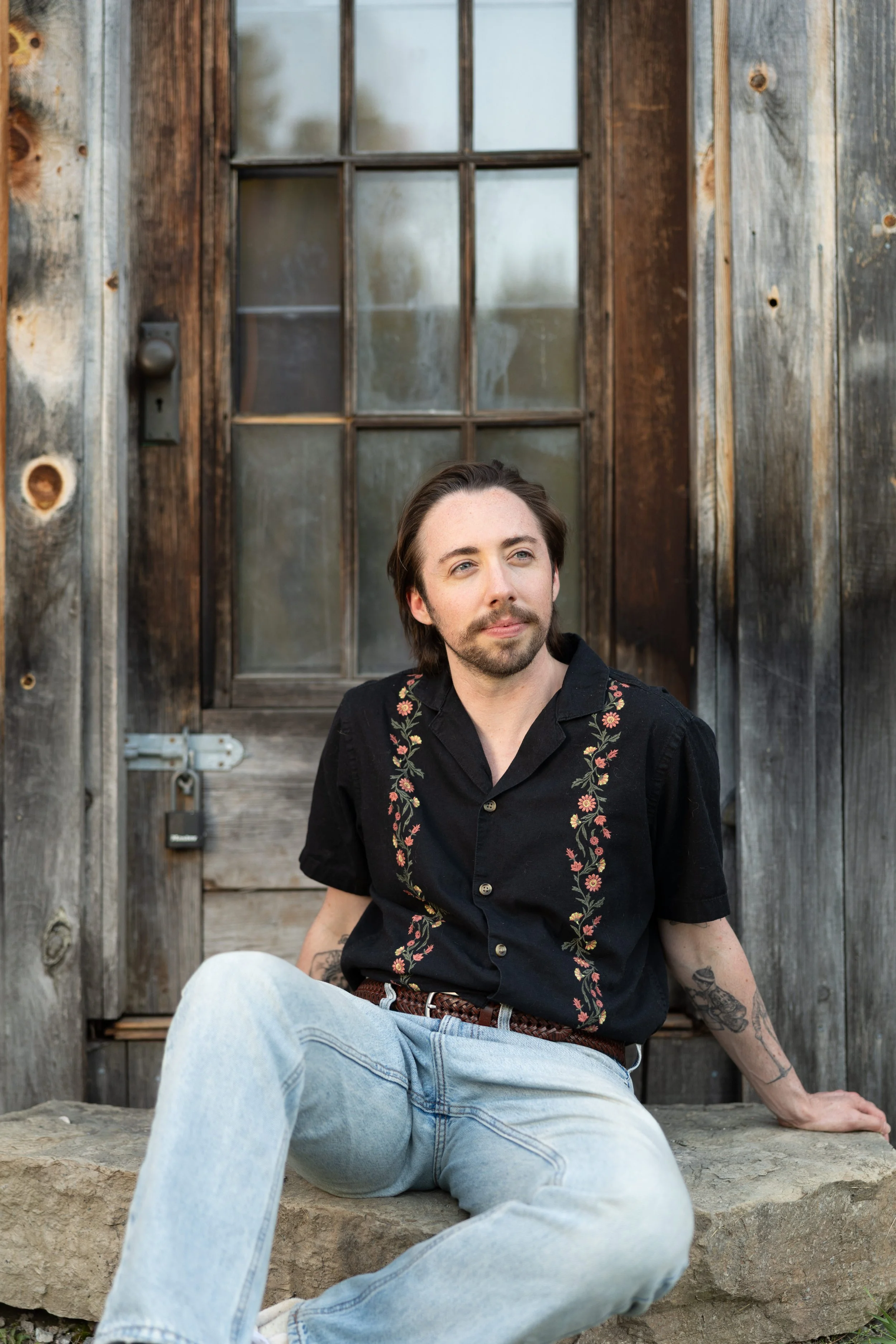 A young man with long hair and facial hair sitting on stone steps in front of a rustic wooden door with glass windows, wearing a black shirt with floral embroidery and light blue jeans.