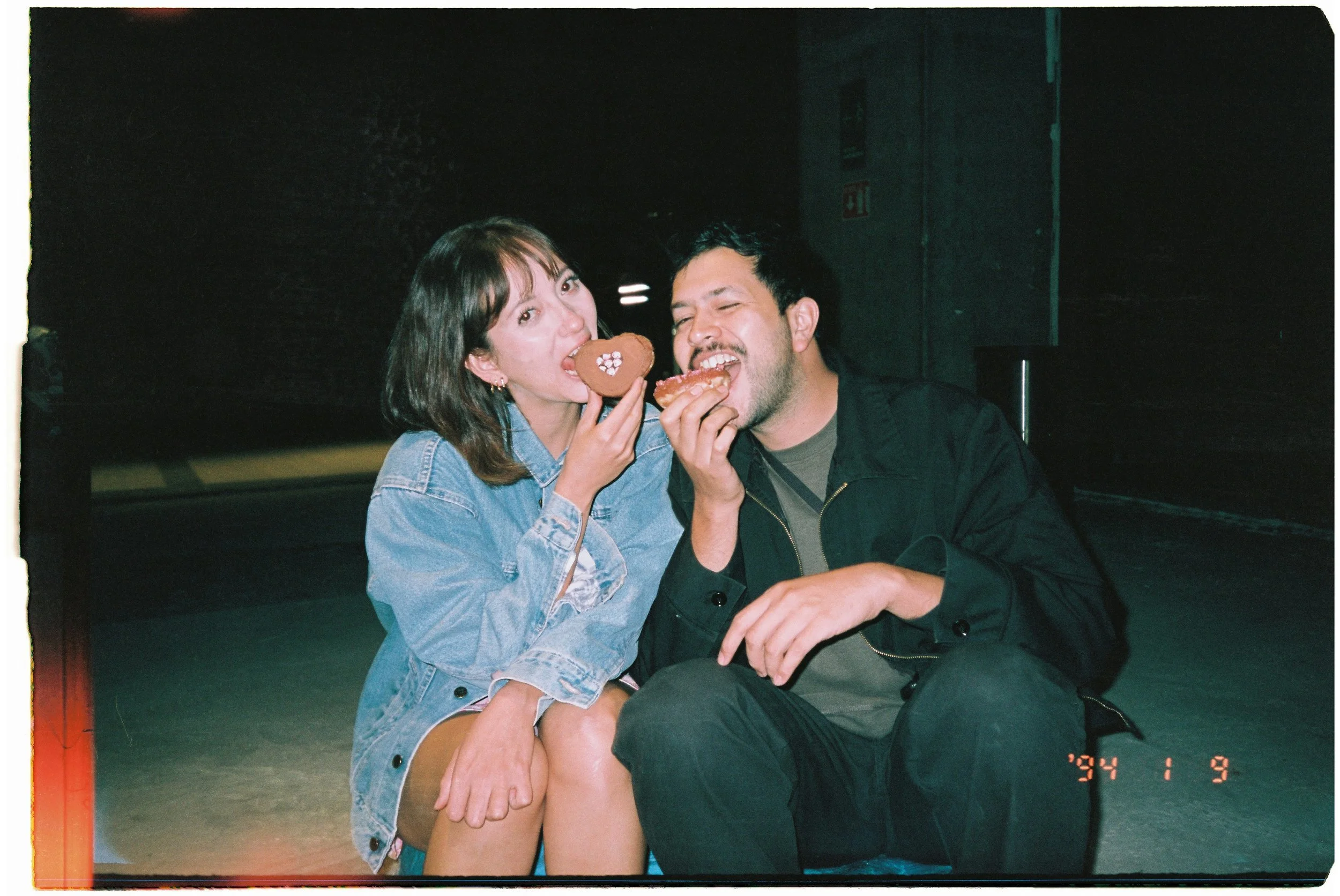 Two people, a woman and a man, sitting together in a dark setting, each holding a heart-shaped pastry with decorative icing, smiling and enjoying their snacks. The woman is wearing a denim jacket, and the man has dark hair and a beard, wearing a black jacket.