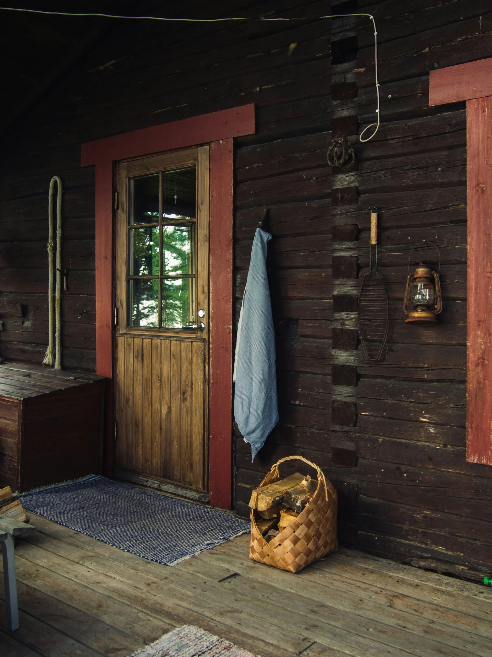 Wooden porch with door, reflection in window, hanging rope, towels, lantern, basket of firewood, and rug.