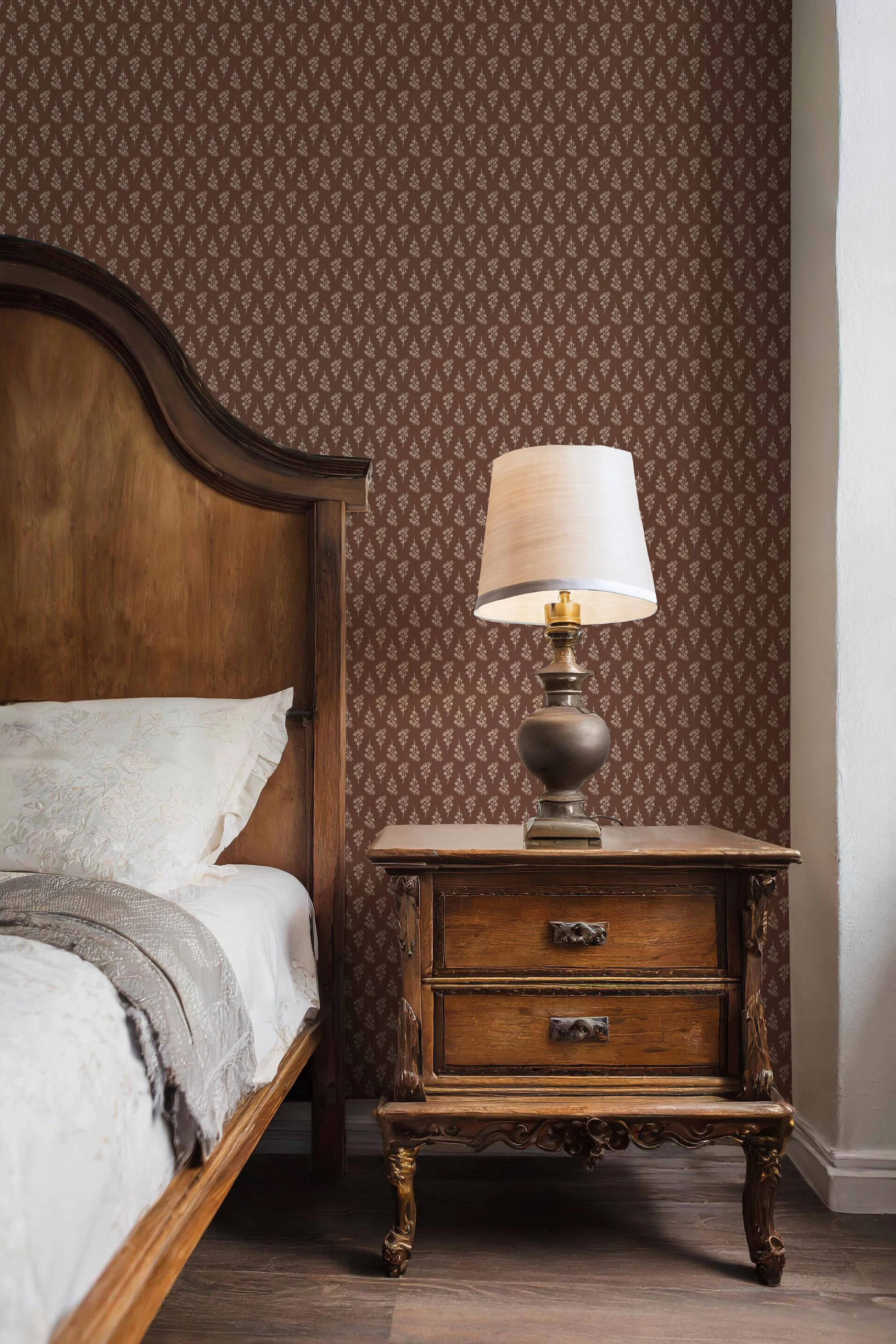 A vintage wooden bedside table with ornate carving, placed next to a bed with white bedding. A lamp with a brass base and white shade sits on the table. The wall behind features dark brown wallpaper with a small floral pattern.