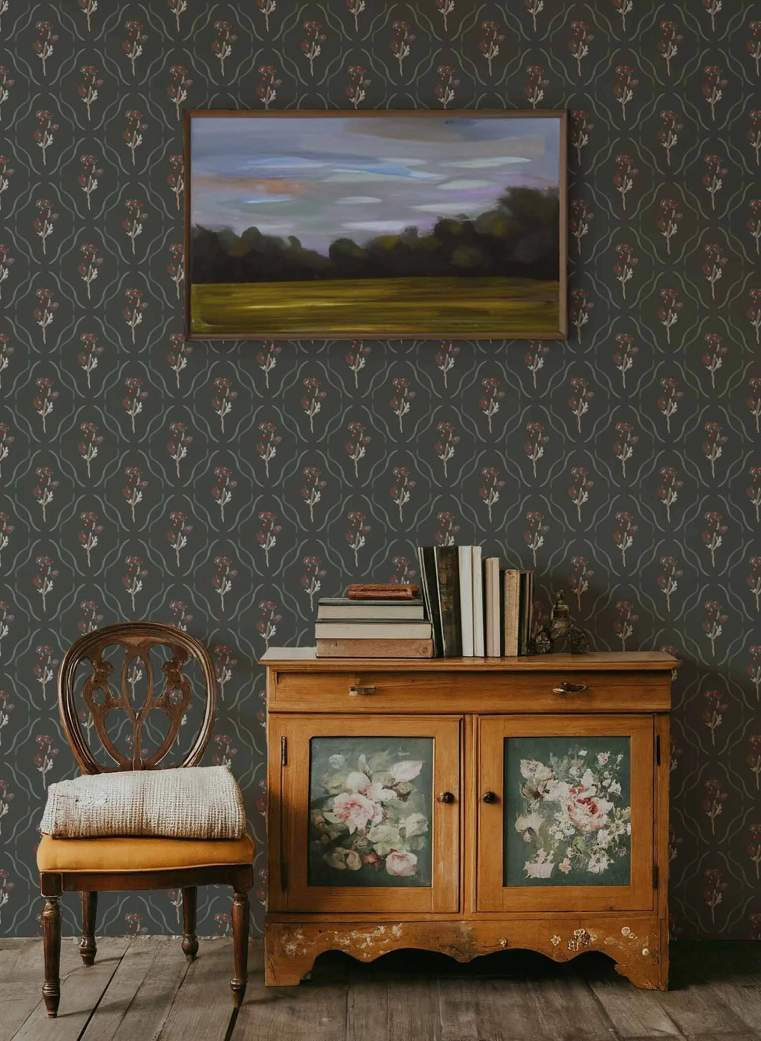 Vintage wooden cabinet with floral paintings on the doors, a stack of books on top, and a wooden chair with a padded woven seat beside it, all set against dark floral wallpaper.