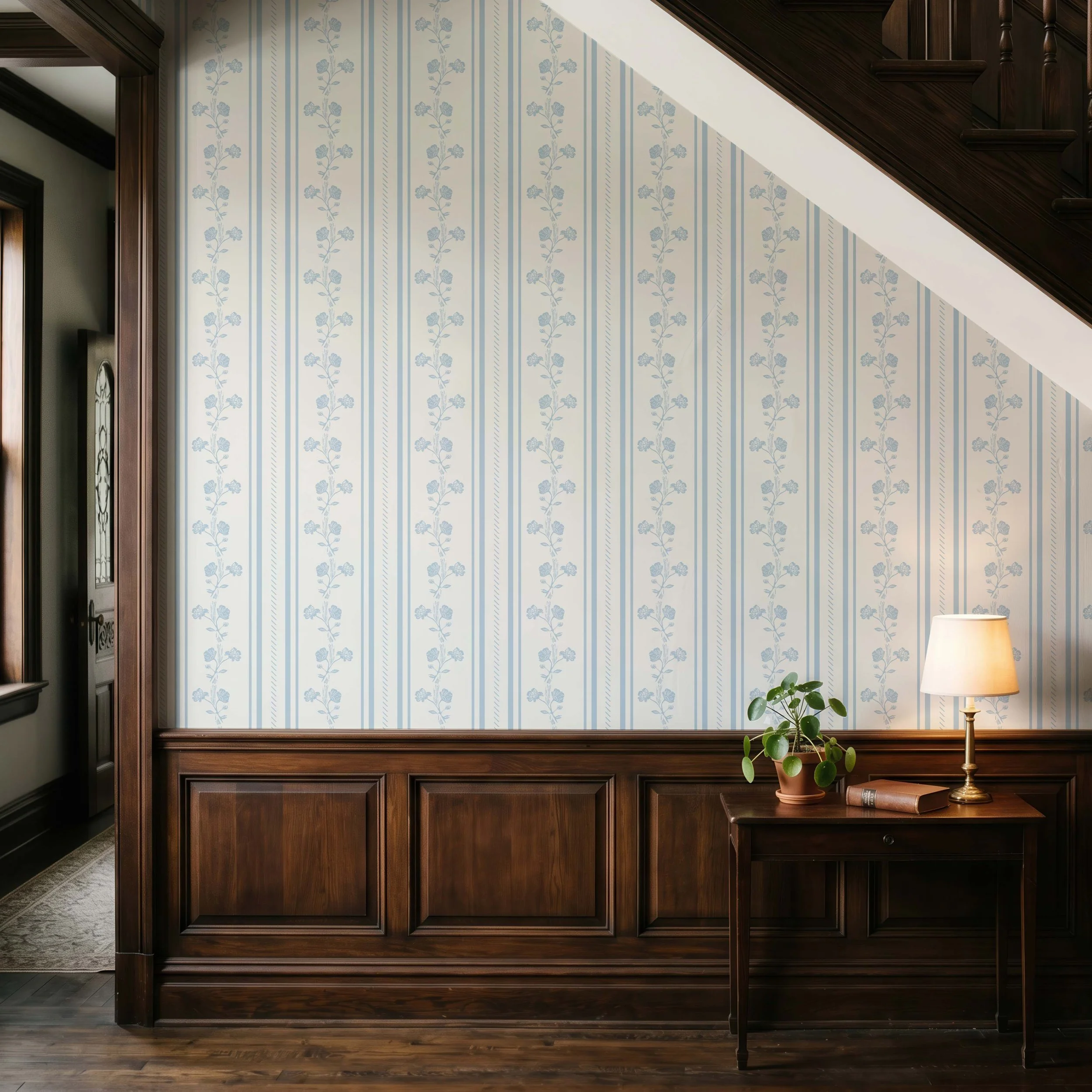 Interior of a room with wood-paneled features and blue floral wallpaper on part of the wall, a table lamp, and a potted plant.