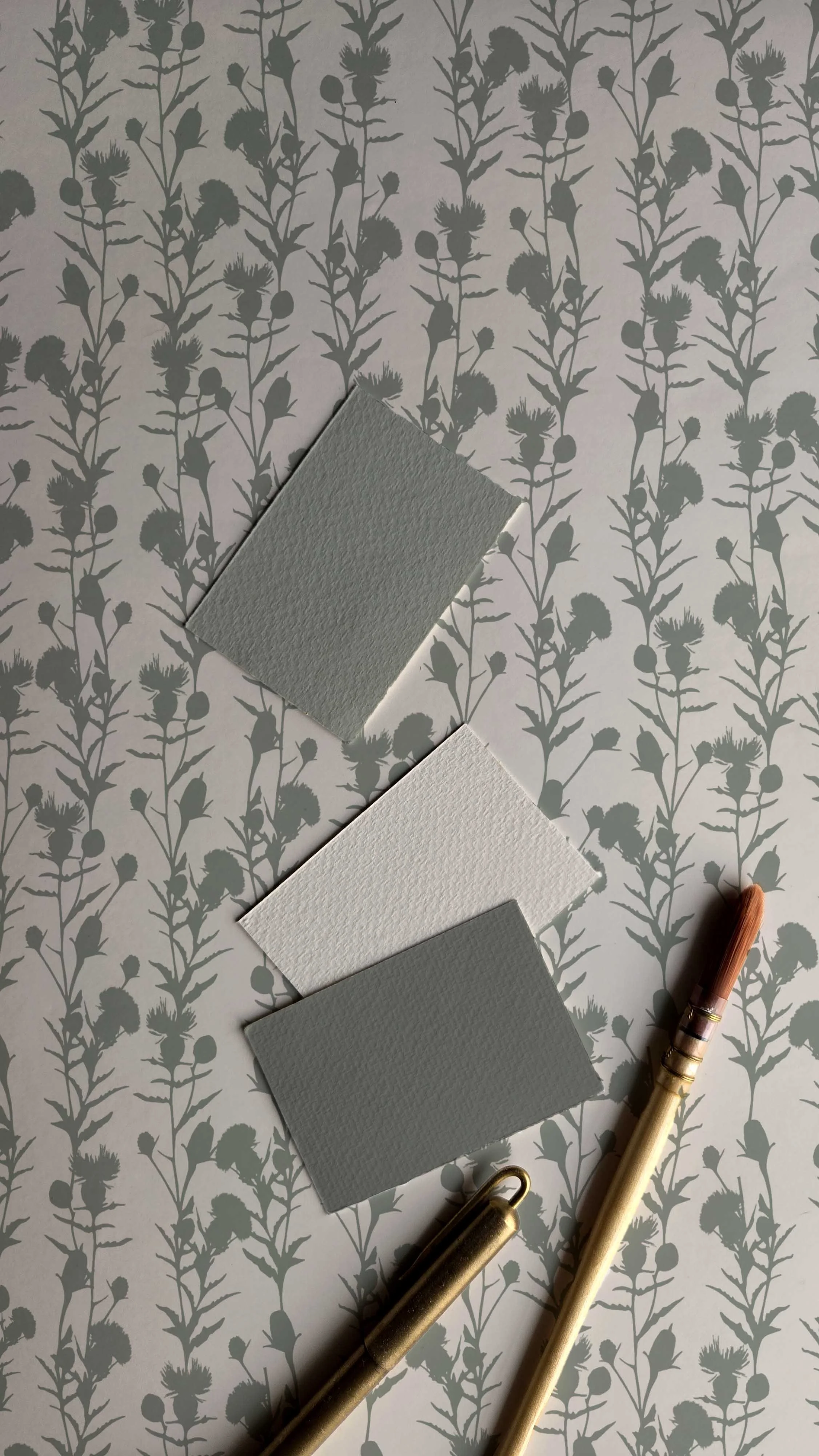 Samples of gray and white textured paper swatches arranged on a gray floral patterned surface with a pen and gold paper clips nearby.